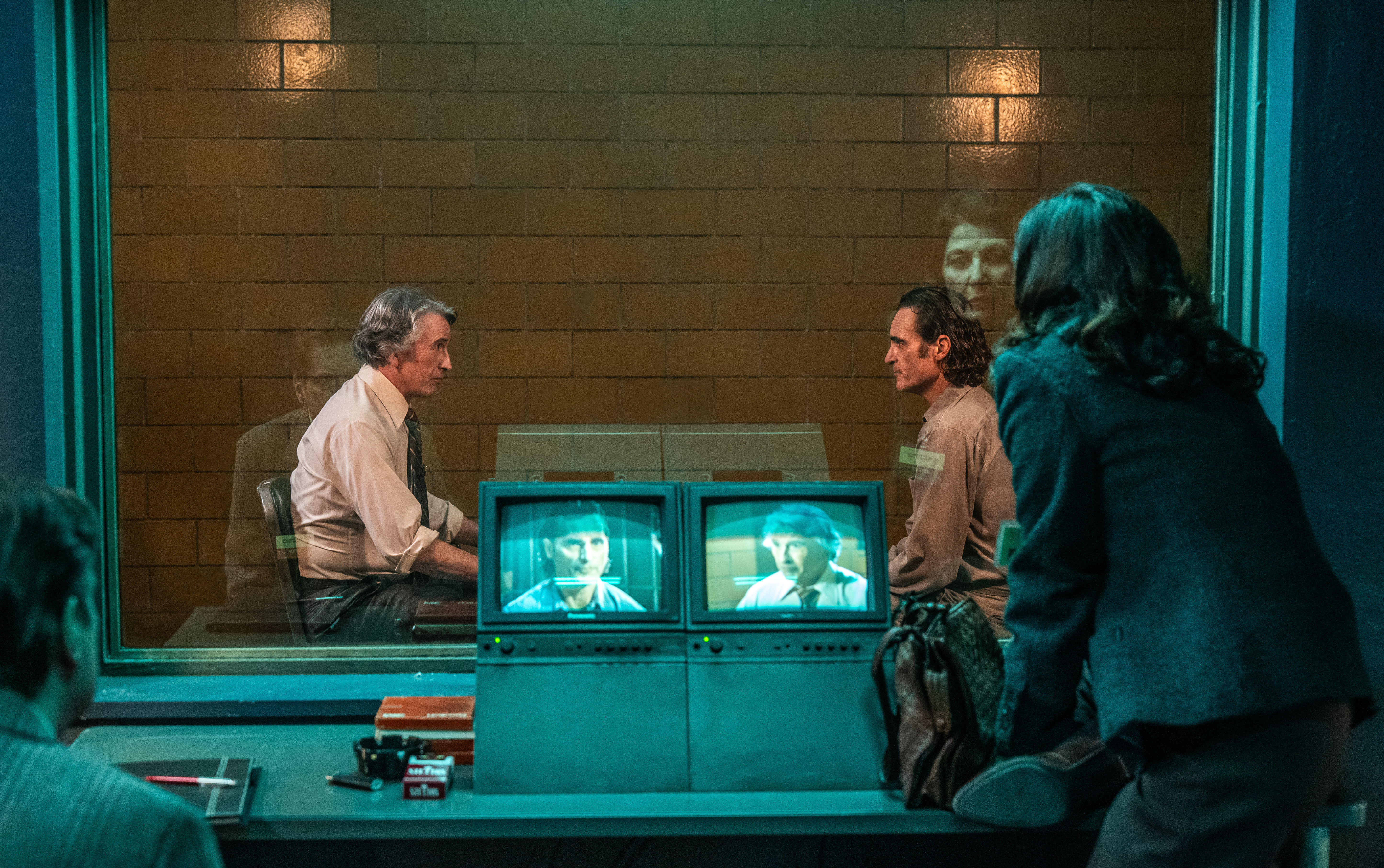 Arthur Fleck (Joaquin Phoenix) and a TV interviewer (Steve Coogan) sit in a barren brick cell in Arkham Asylum, facing each other for an interview, as others watch them on monitors in Joker: Folie à Deux