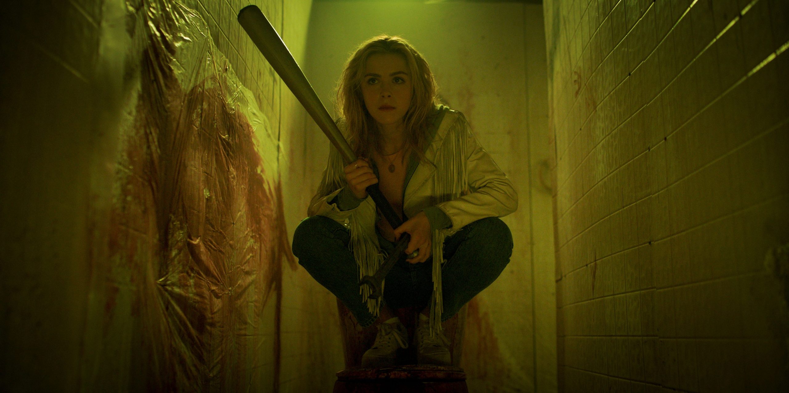 Kiernan Shipka hunched on top of a toilet under a sickly yellow light holding a baseball bat in Totally Killer.