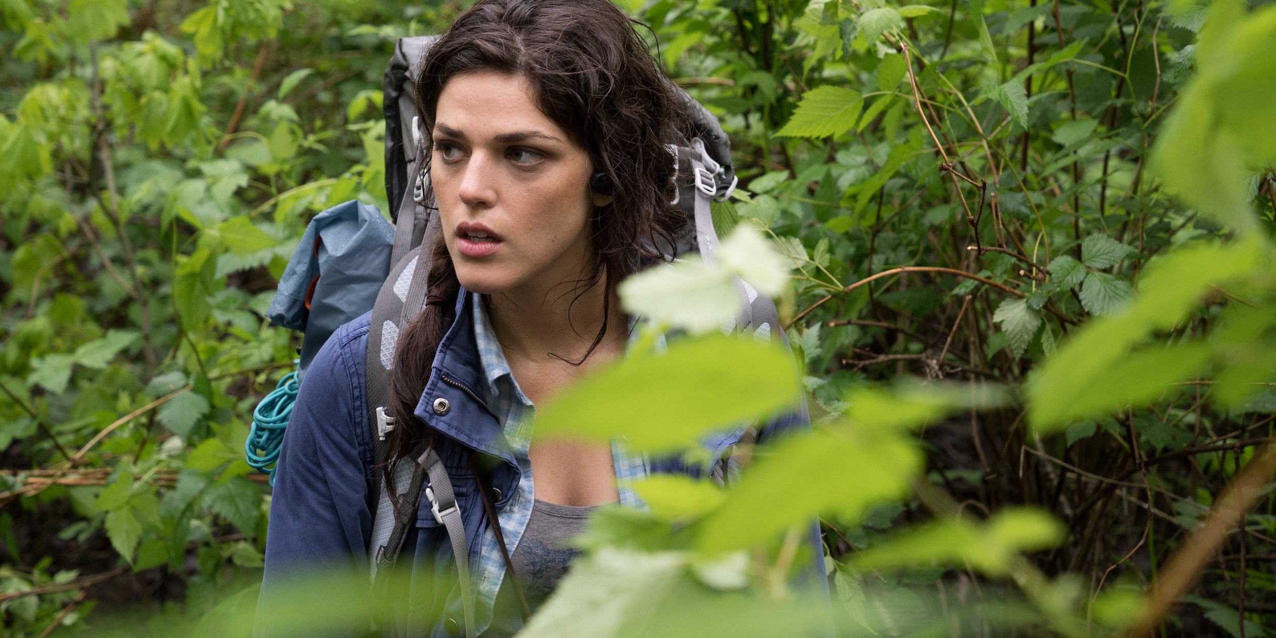 A woman stands behind bushes with a backpack on in the 2016 Blair Witch movie