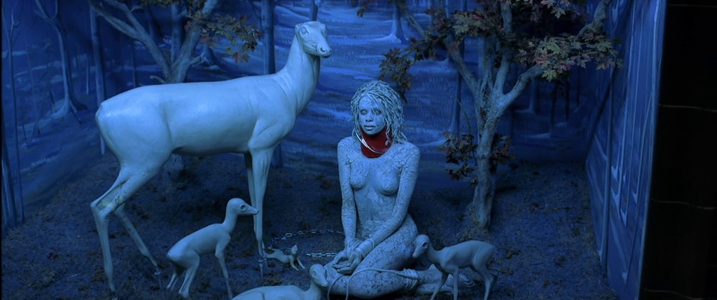 A woman covered in white makeup and bound in a menagerie surrounded by statues of deer and fawn in The Cell (2000).