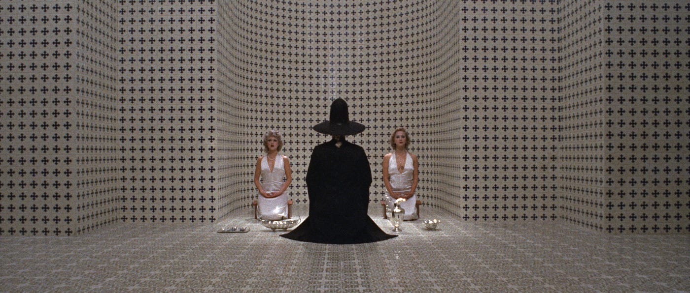 A person in a black cloak and hat kneeling in the middle of a checkered patterned room flanked by women in white outfits in The Holy Mountain.