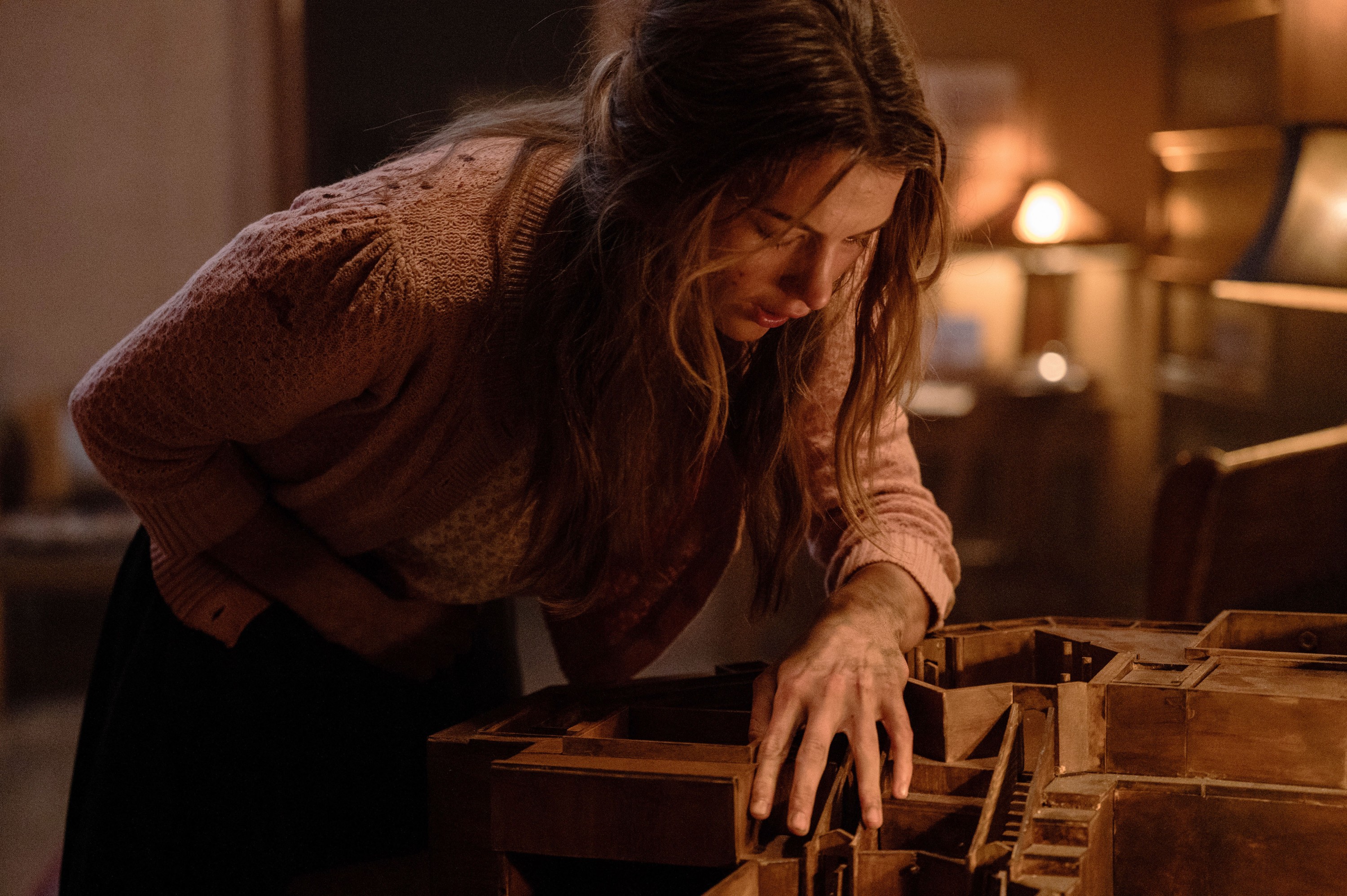 Sister Paxton (Chloe East, wearing a pink sweater, with her hair dangling around her face) clutches her stomach as she leans over a small wooden model of the house she’s trapped in in the horror movie Heretic