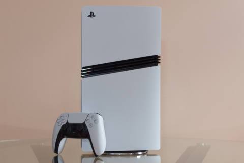 A photo of the PlayStation 5 Pro console resting on a glass table, set against a peach-colored wall. The DualSense controller is leaning against the console.