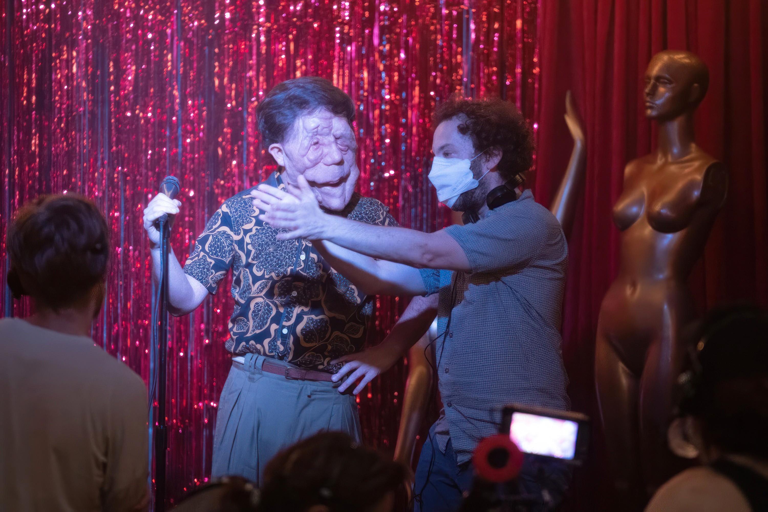 Actor Adam Pearson stands at a karaoke mic, as director Aaron Schimberg, in a white surgical mask holds up his hands on the set of A Different Man