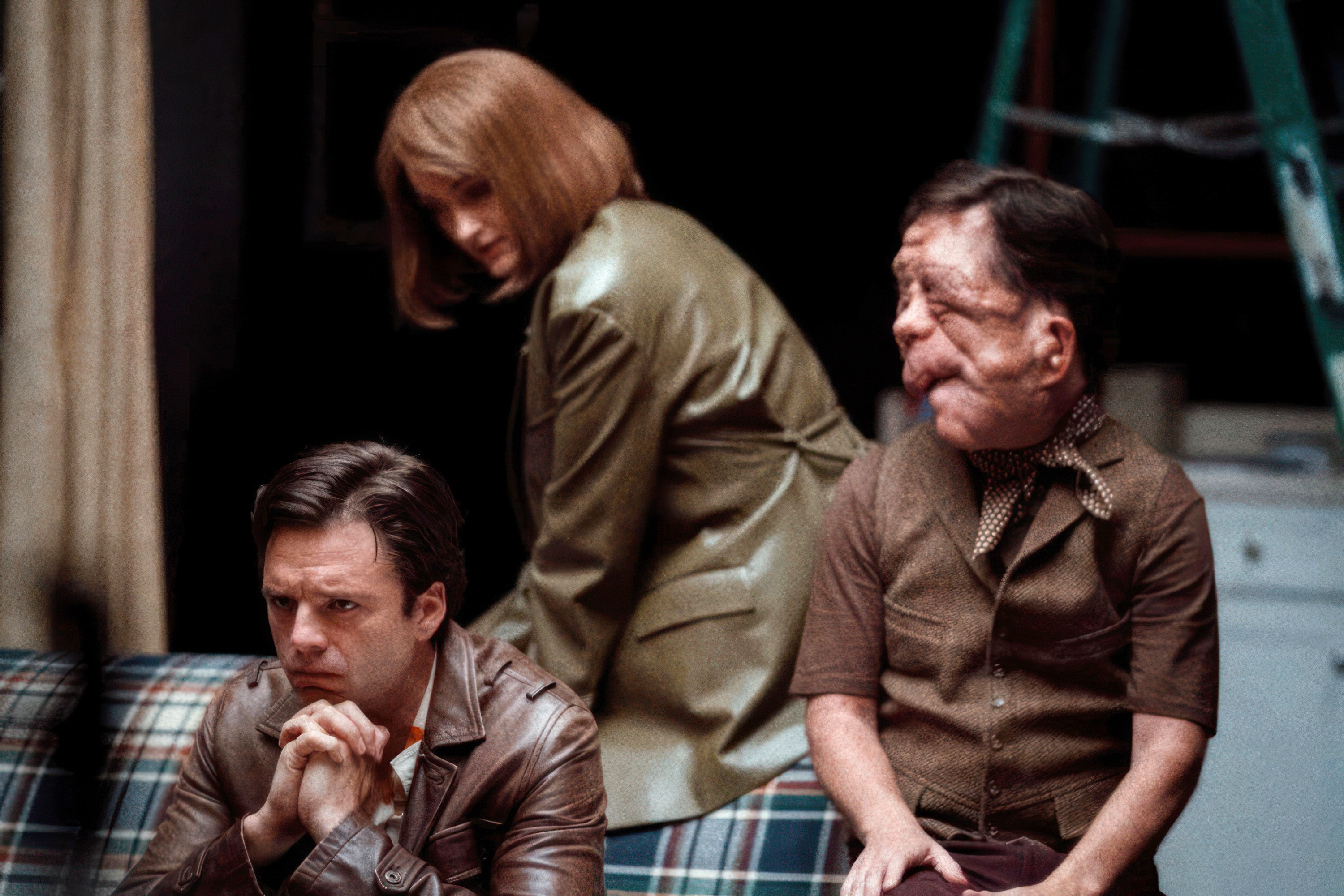 Edward (Sebastian Stan) sits pensively on a couch on a stripped-down theater stage as Ingrid (Renate Reinsve) and Oswald (Adam Pearson) attempt to comfort him in A Different Man