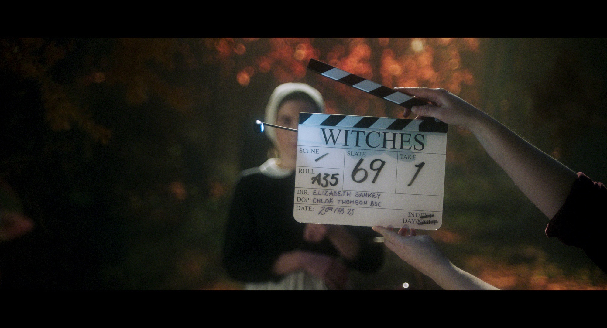 A pair holding a clapperboard with the word “Witches” written on it in front of a woman standing in an autumnal forest dressed in a puritan gown in Witches.