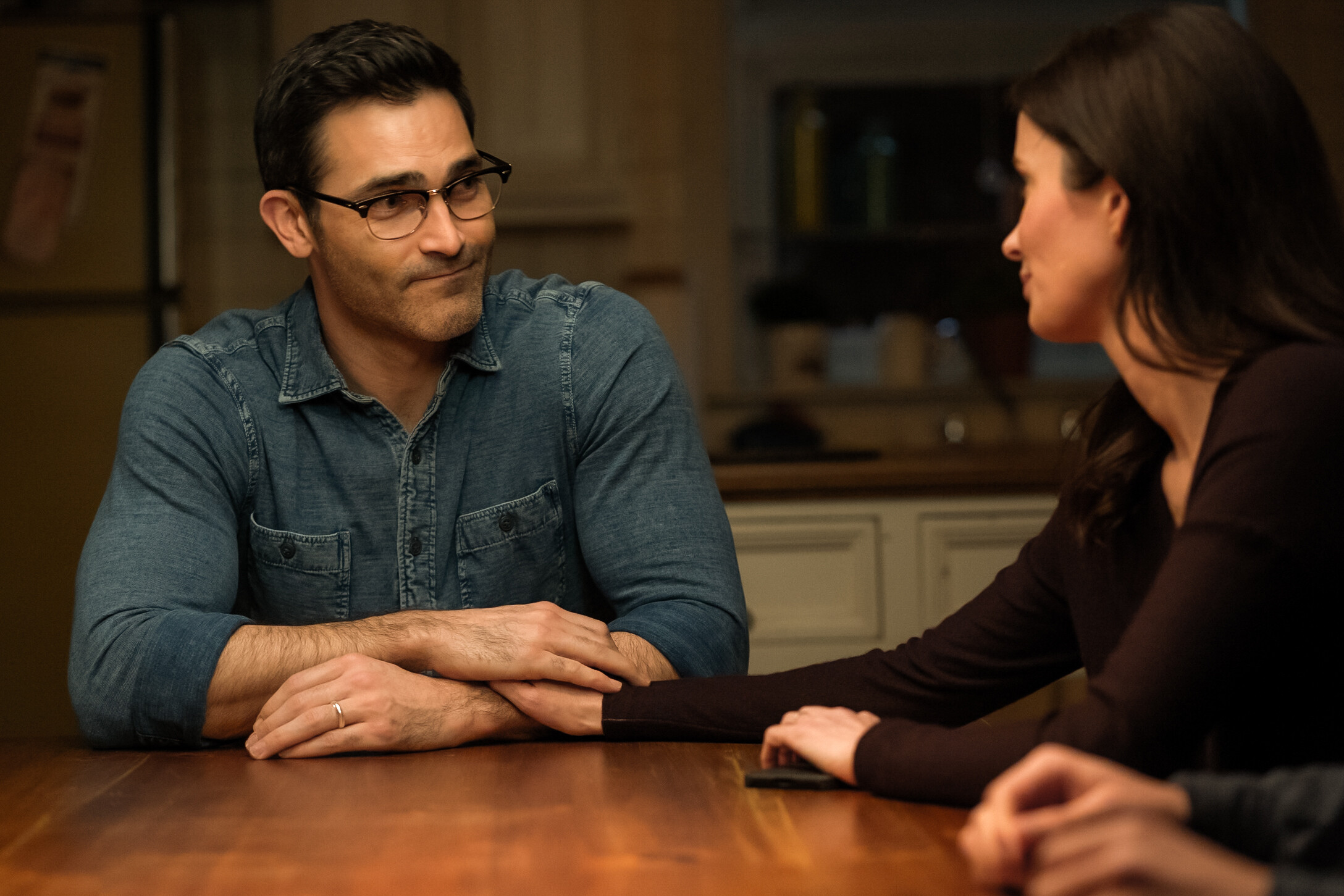 Clark (Tyler Hoechlin) sitting at the kitchen table with Lois (Elizabeth Tulloch) talking and holding hands, in a still from Superman & Lois season 4