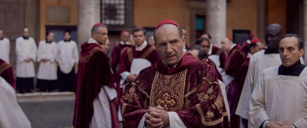 Ralph Fiennes dressed in clergical robes standing in a room filled with clergymen in Conclave.