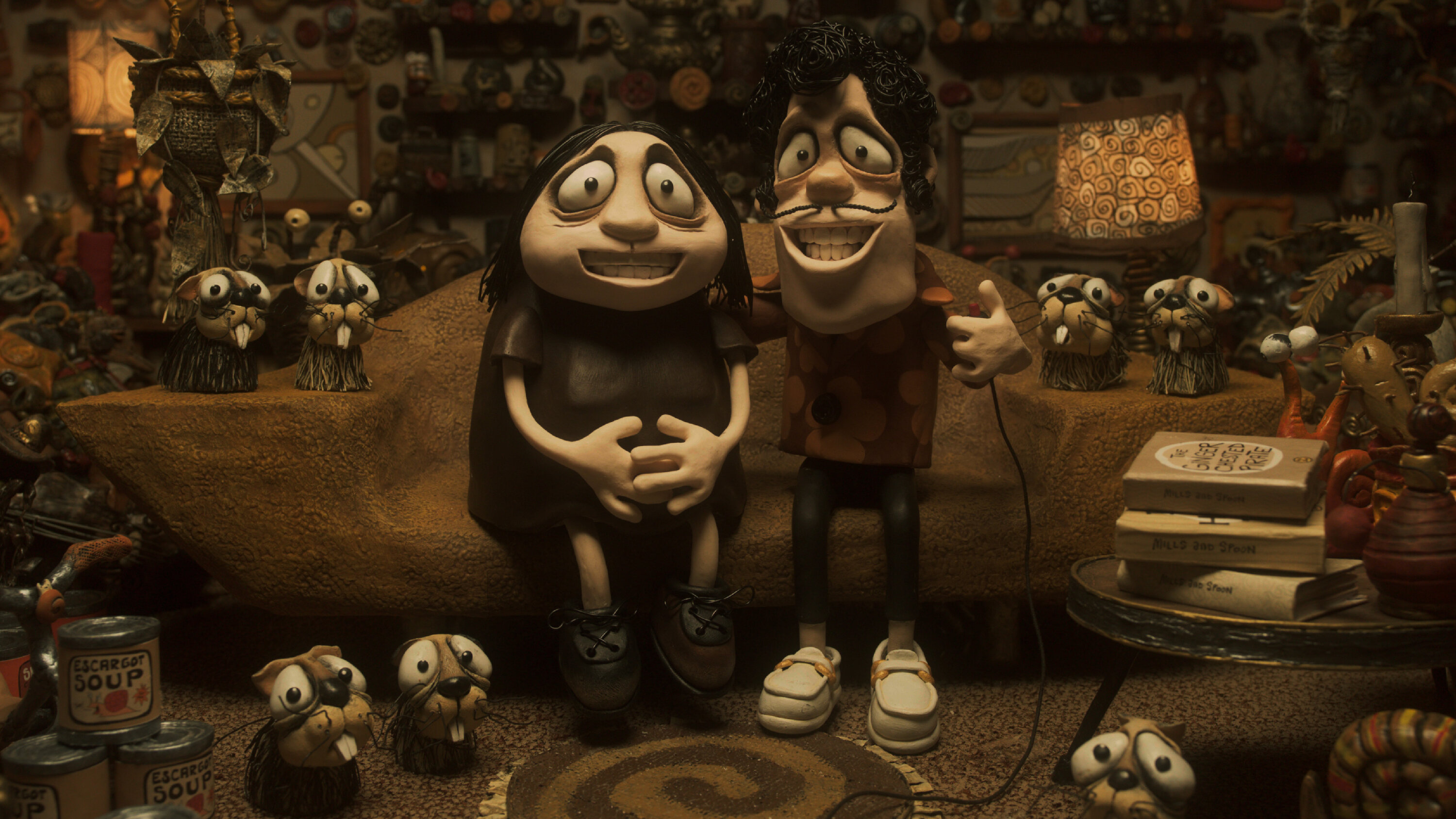 Two claymation figures smiling and sitting in a living room cluttered with adorable creatures and ephemera in Memoir of the Snail.