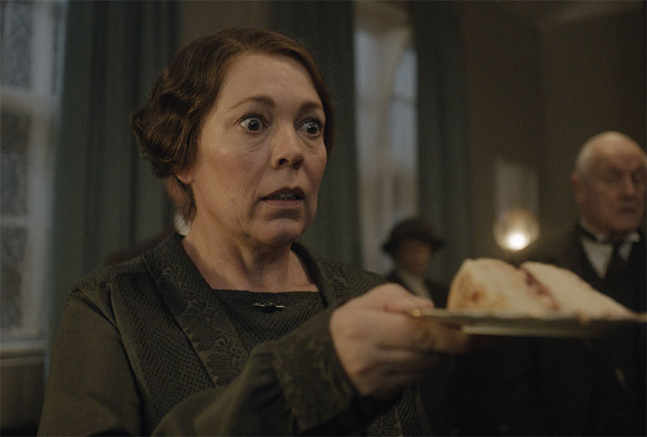 Olivia Colman, surprised, holds a slice of cake on a plate in Wicked Little Letters