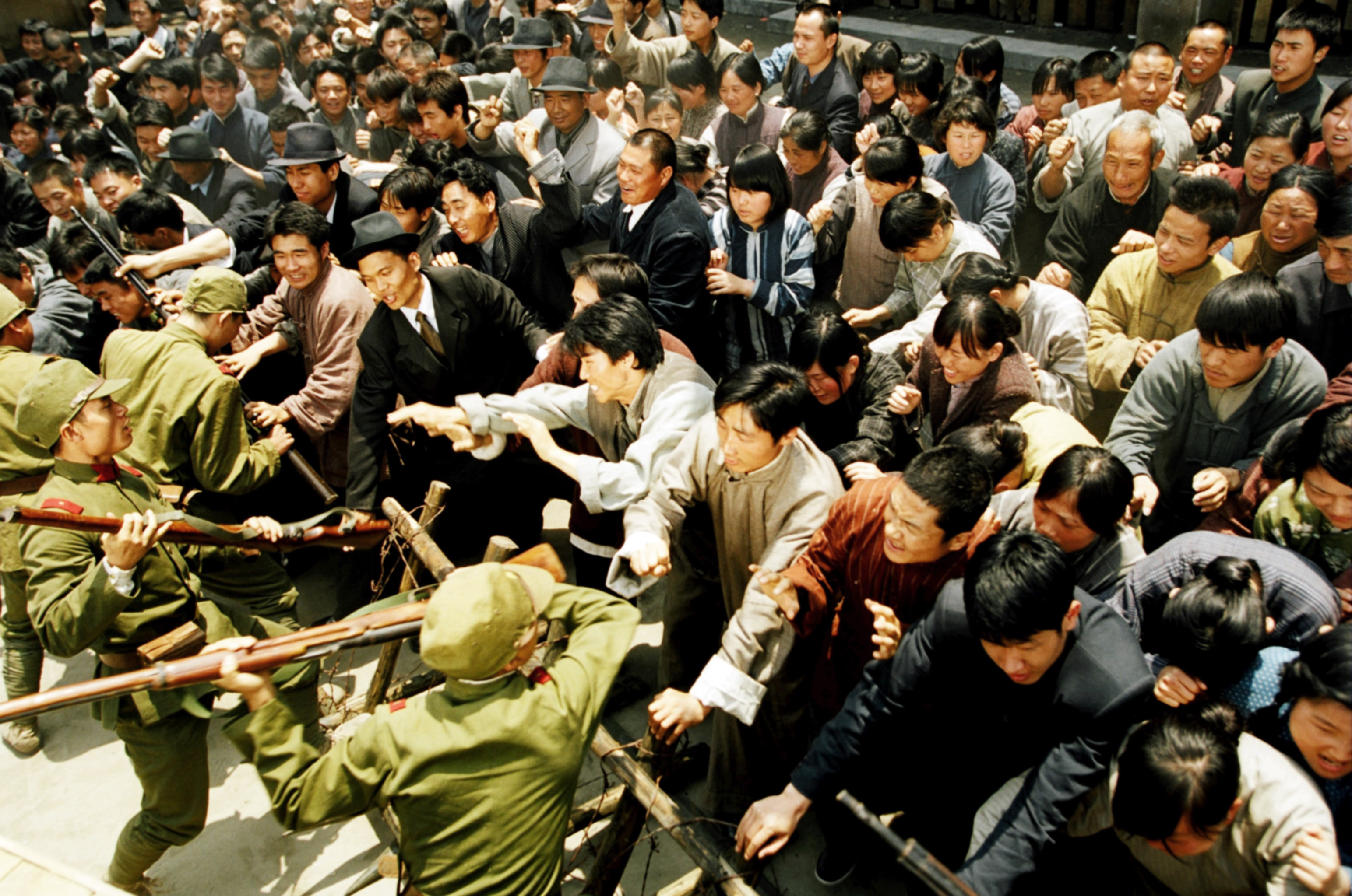 A crowd of civilians in black, white, and brown surges against a barricade manned by soldiers in green in a scene from 2008’s Ip Man