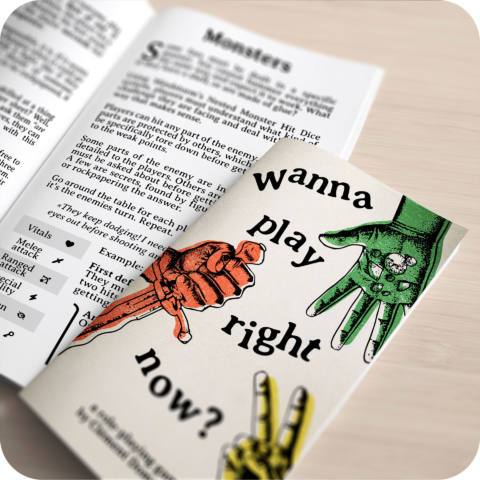 A picture of the book for “wanna play right now” on a table, with the cover depicting three hands, one green with an open palm, one red with a knife, one yellow with two fingers out, mirroring “rock paper scissors”