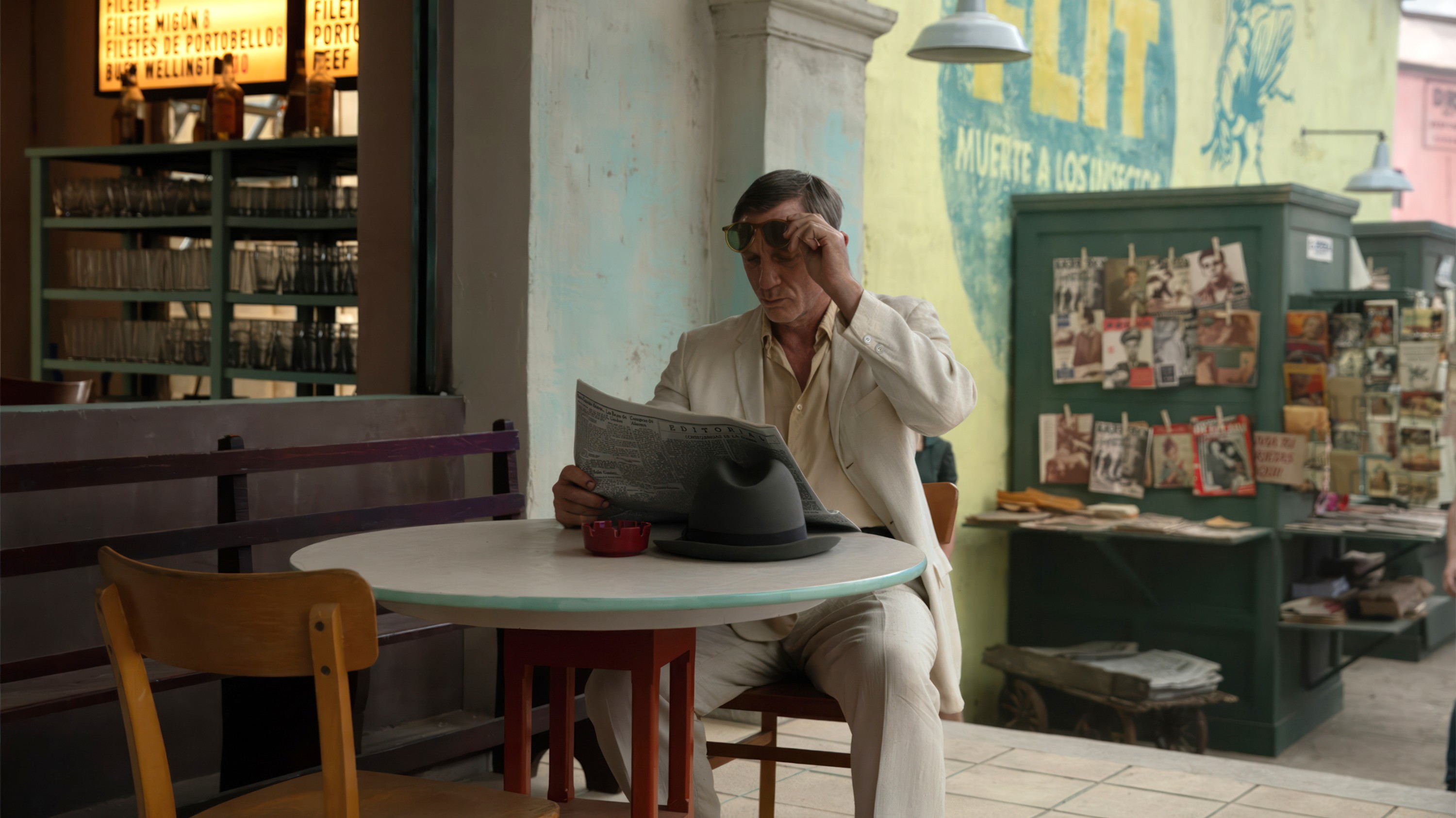 In Queer, Daniel Craig, wearing a nice summer suit, sits at a coffee table outside a restaurant and reads the newspaper