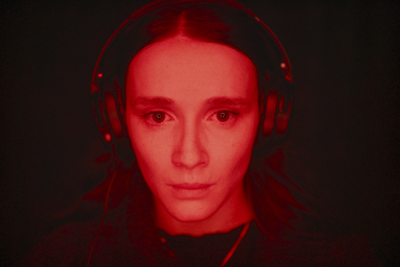 Juliette Gariépy looks toward the camera while wearing headphones and bathed in red light in Red Rooms Juliette Gariépy looks toward the camera while wearing headphones and bathed in red light in Red Rooms