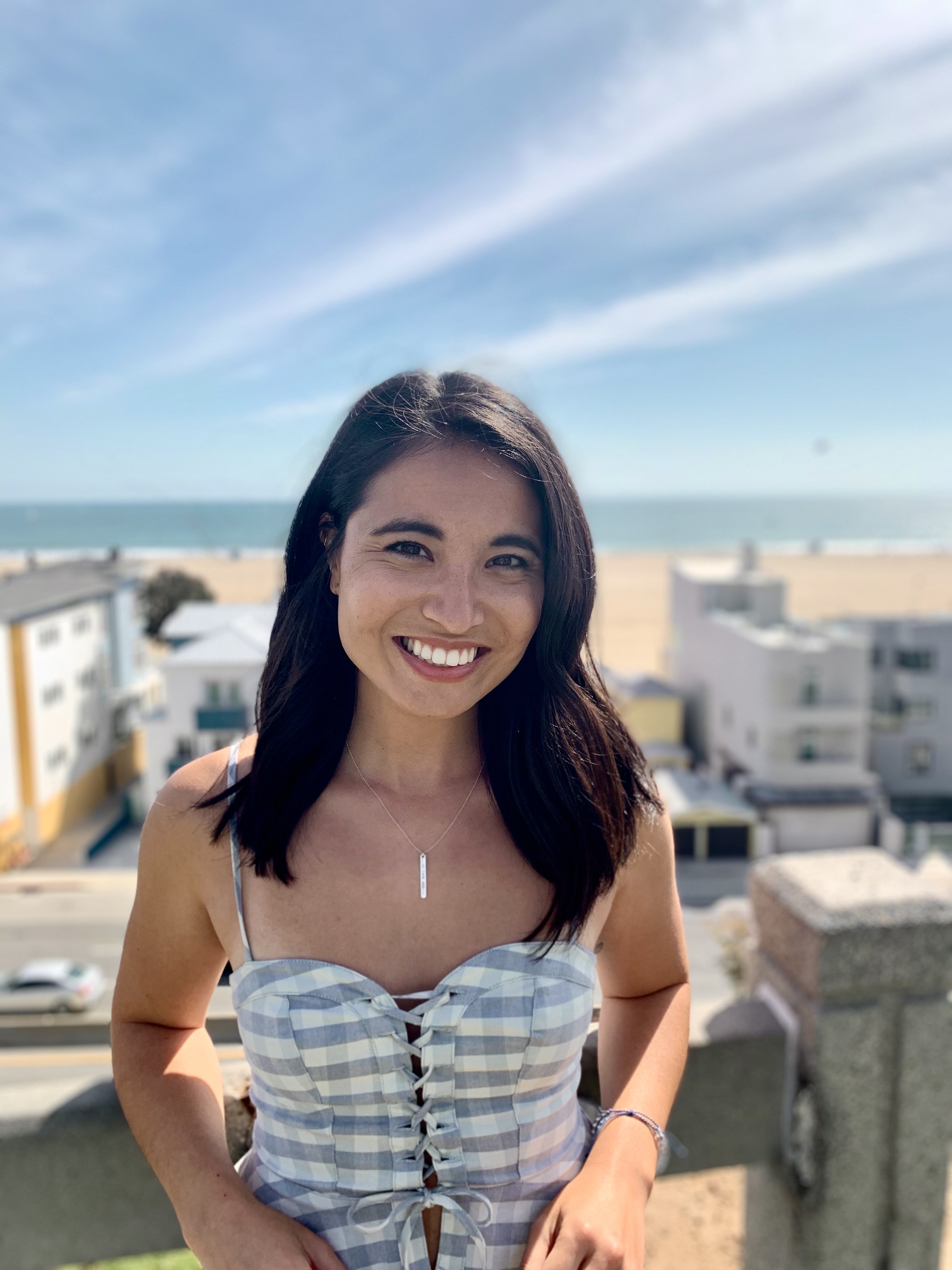 Author Olivie Blake standing on a roof smiling at the camera, with buildings and a beach behind her in the background