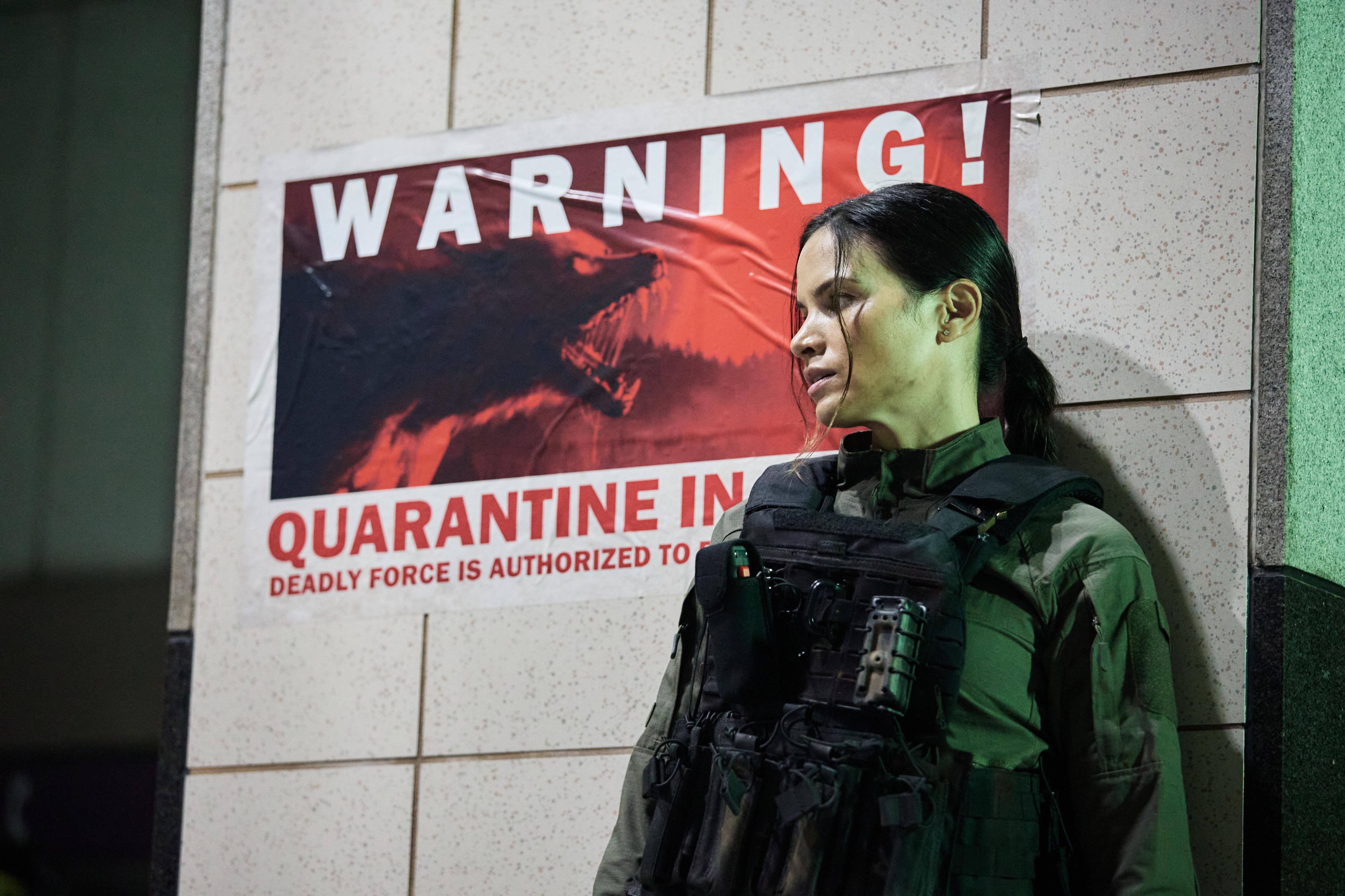 Katrina Law, wearing a bulletproof vest, stands against a wall featuring a poster warning about quarantine due to werewolves, in Werewolves