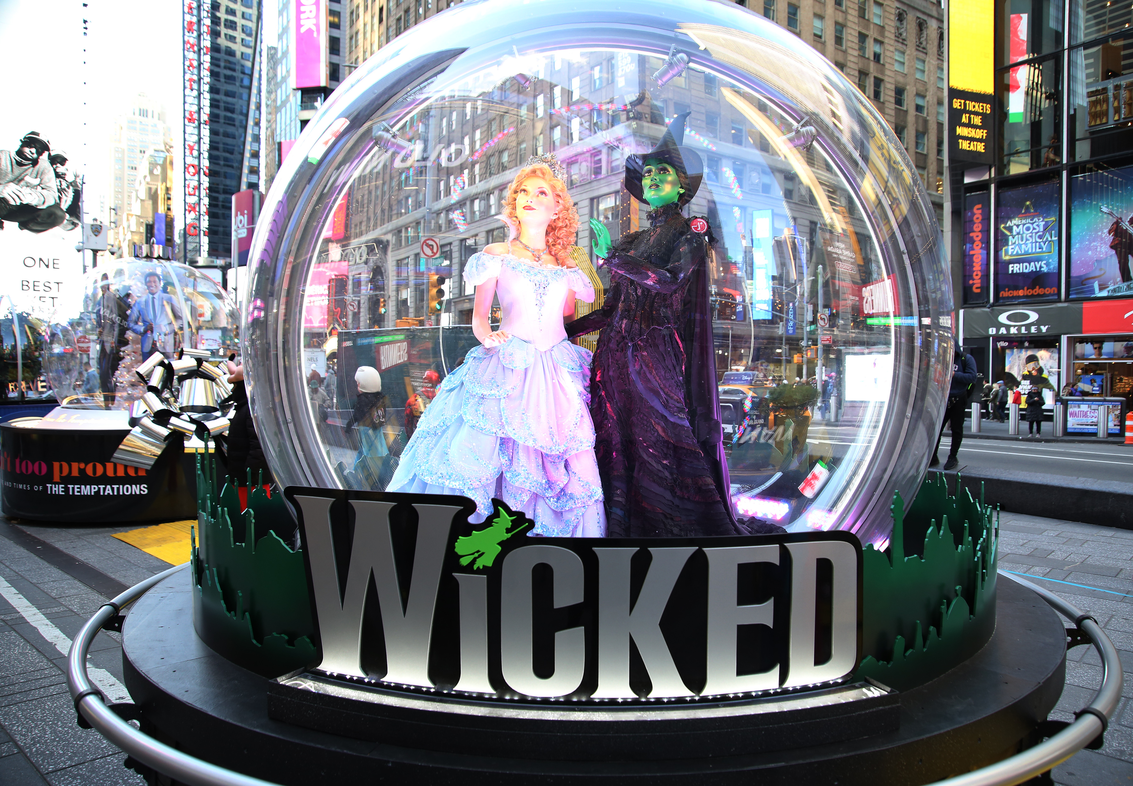 A snow globe featuring statues of Glinda and Elphaba standing together stands in Times Square, with “WICKED” on it in huge letters