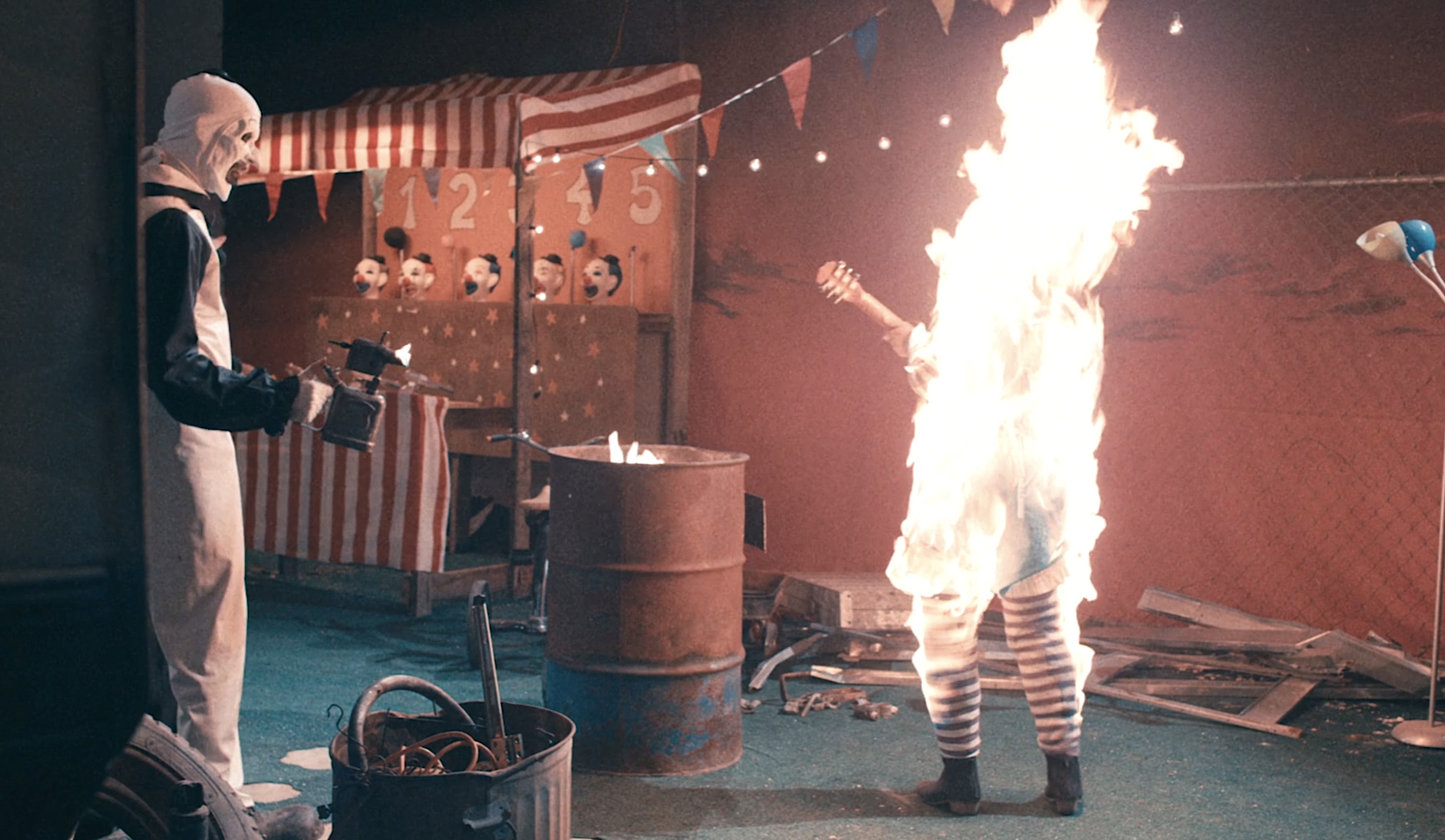 Art the Clown stands in a run-down clown café, holding a flamethrower, while a female clown in striped stockings, playing a guitar, disappears under a whoosh of fire in a sequence from Terrifier 2 Art the Clown stands in a run-down clown café, holding a flamethrower, while a female clown in striped stockings, playing a guitar, disappears under a whoosh of fire in a sequence from Terrifier 2
