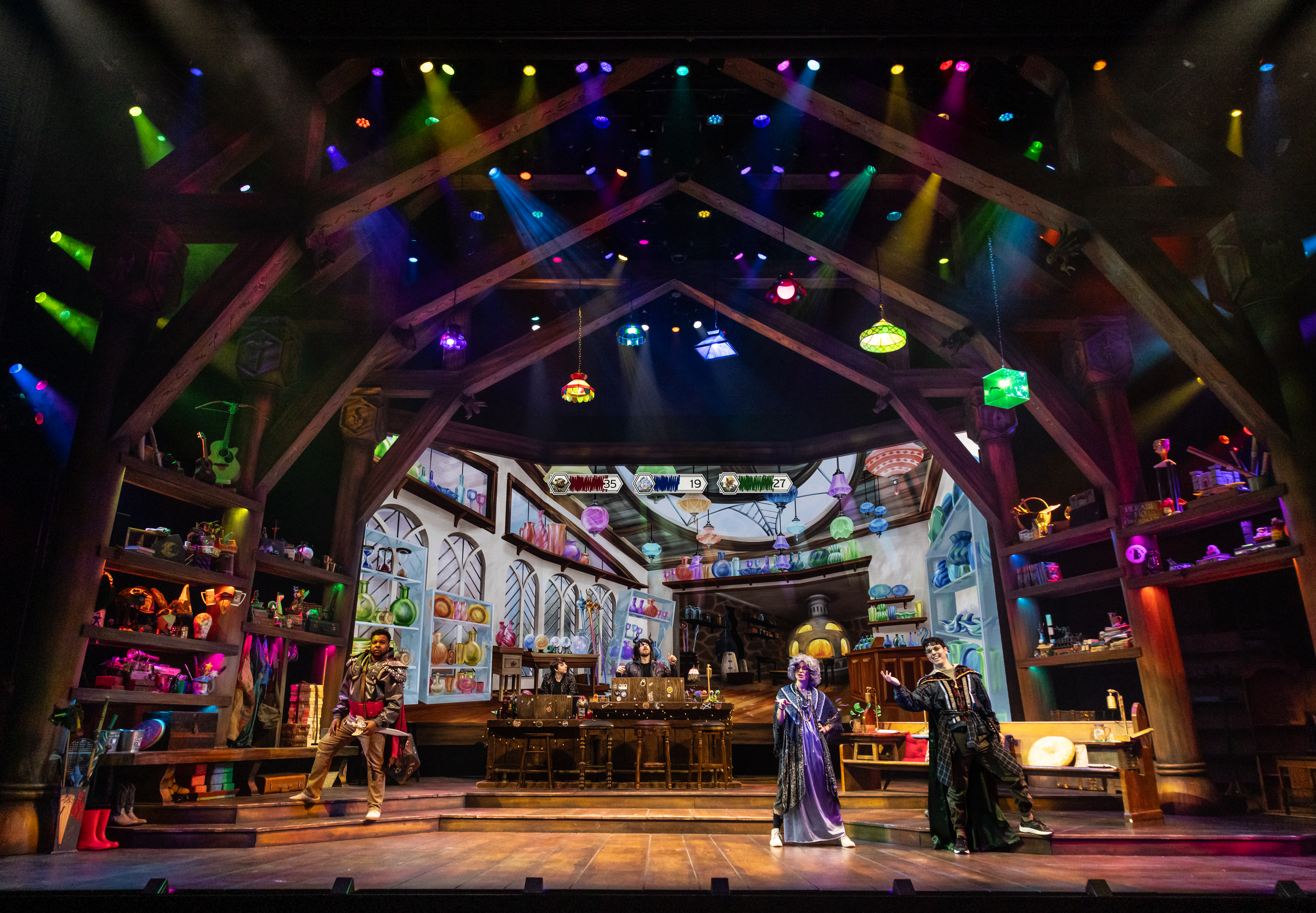 The stage of Twenty-Sided Tavern, with a large projection screen, hanging lights, props on the wall, and performers in costume The stage of Twenty-Sided Tavern, with a large projection screen, hanging lights, props on the wall, and performers in costume