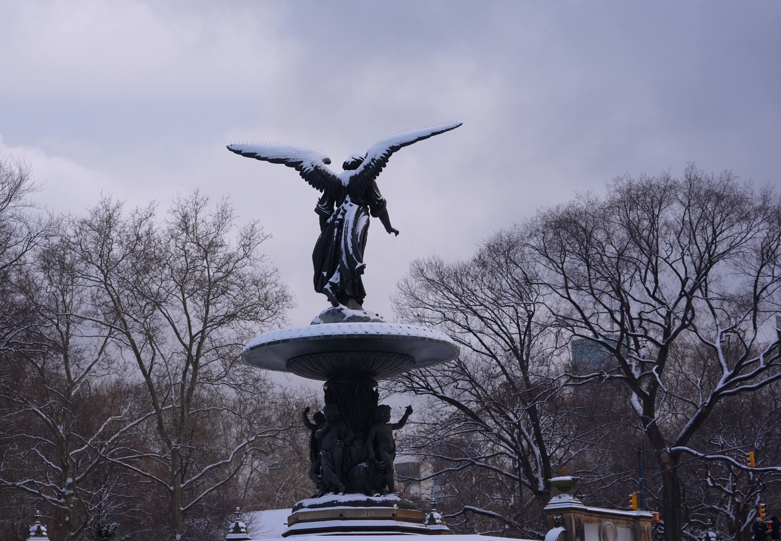 A snow-covered statue of NYC’s Bethesda Terrace’s Angel of the Waters statue atop a sculpted plinth. Behind it are grey winter skies and bare treetops.