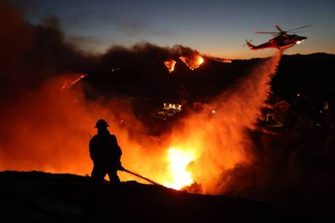 Fire personnel respond to homes destroyed while a helicopter drops water as the Palisades Fire grows in Pacific Palisades, California on January 7, 2025. A fast-moving wildfire in a Los Angeles suburb burned buildings and sparked panic, with thousands ordered to evacuate January 7, 2025 as “life threatening” winds whipped the region. Frightened residents abandoned their cars on one of the only roads in and out of the upscale Pacific Palisades area, fleeing on foot from the 770-acre (310-hectare) blaze engulfing an area crammed with multi-million dollar homes in the Santa Monica Mountains.