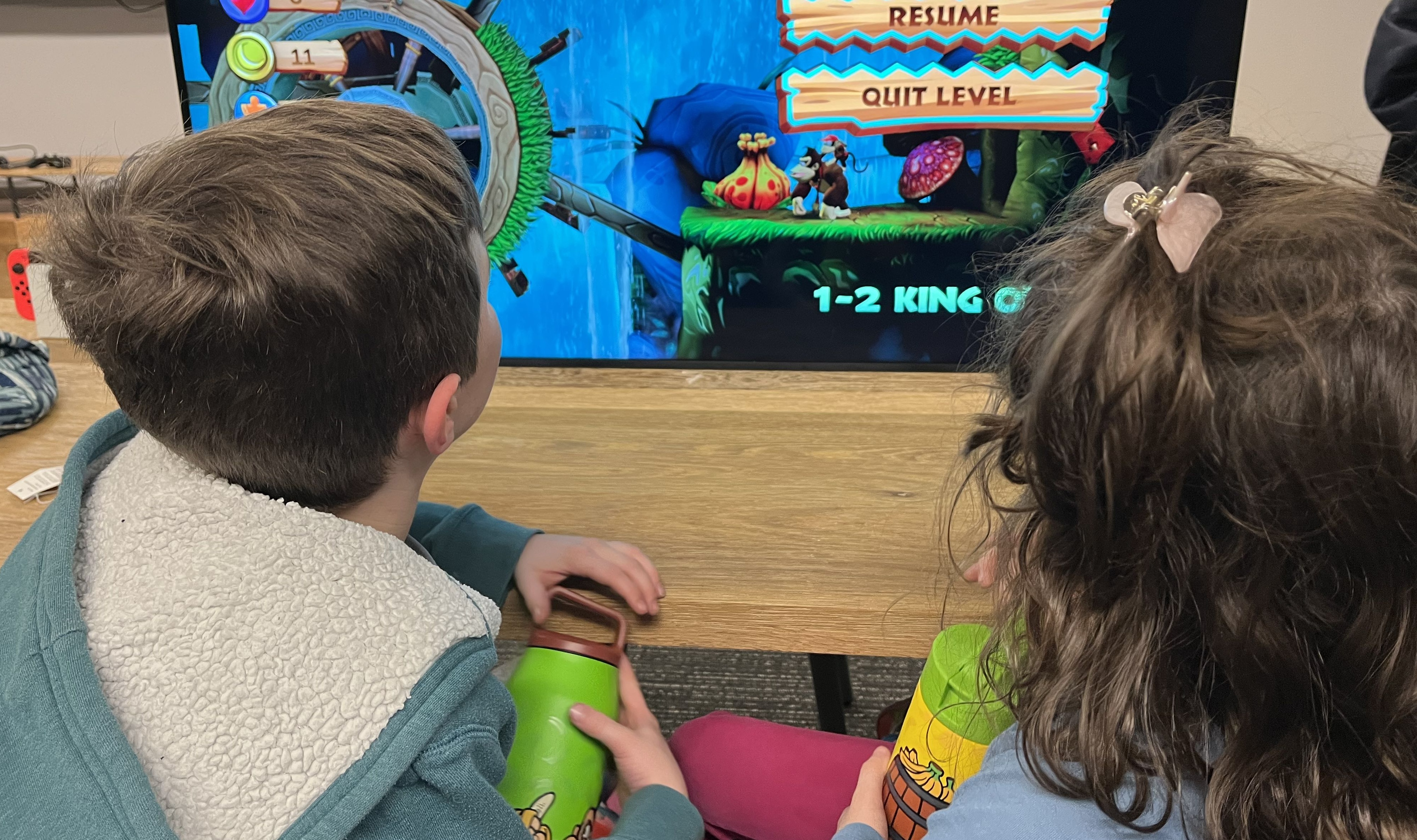 Two children facing away from the viewer, towards a large TV screen depicting Donkey Kong Country Returns HD Two children facing away from the viewer, towards a large TV screen depicting Donkey Kong Country Returns HD