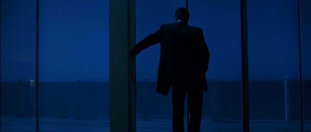 A man in a suit standing next to a window overlooking a beach at night in Heat.