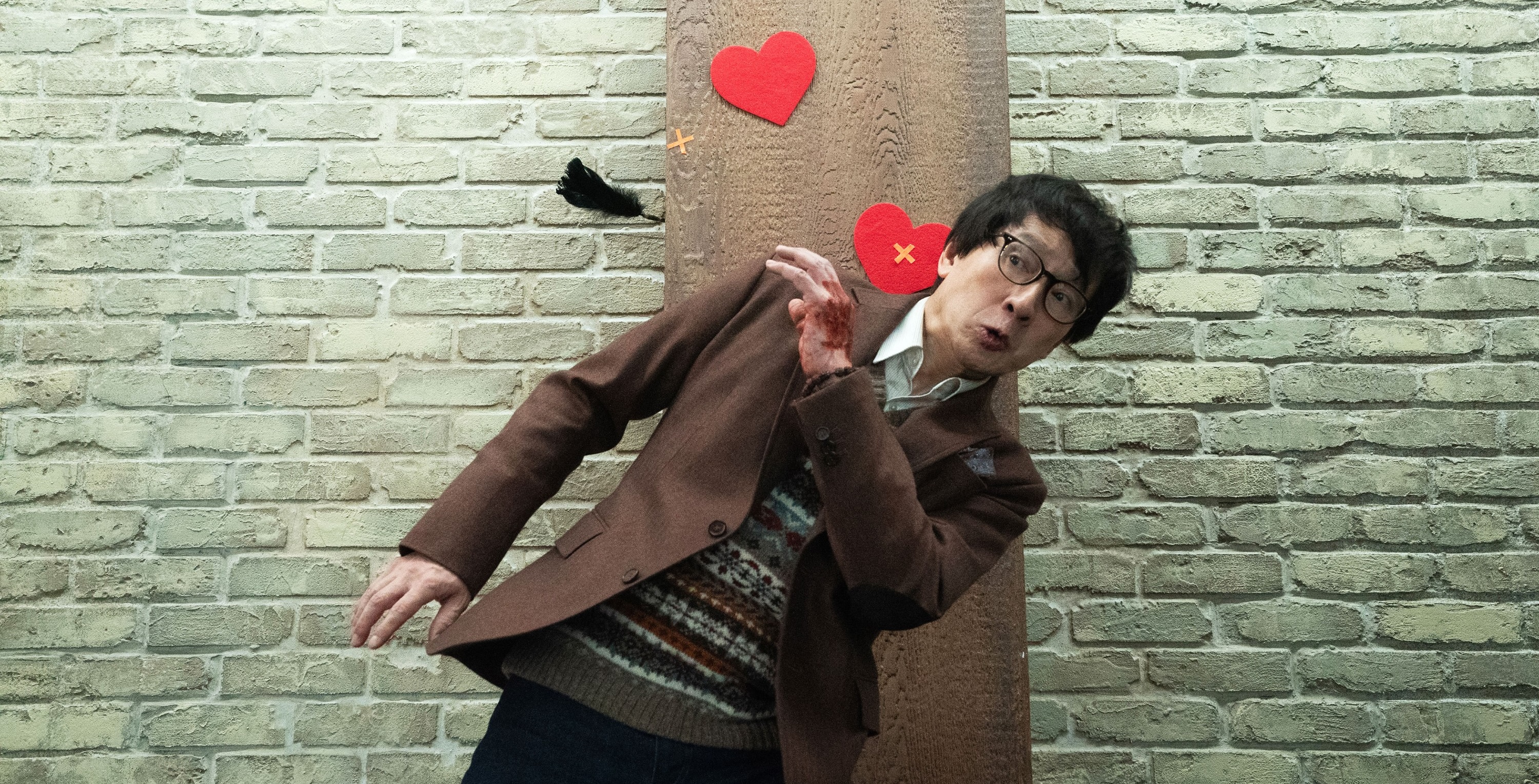 Marvin (Ke Huy Quan), stands in front of a brick wall dotted with paper heart cutouts and dodges a flying black feather with a comically exaggerated expression on his face in Love Hurts