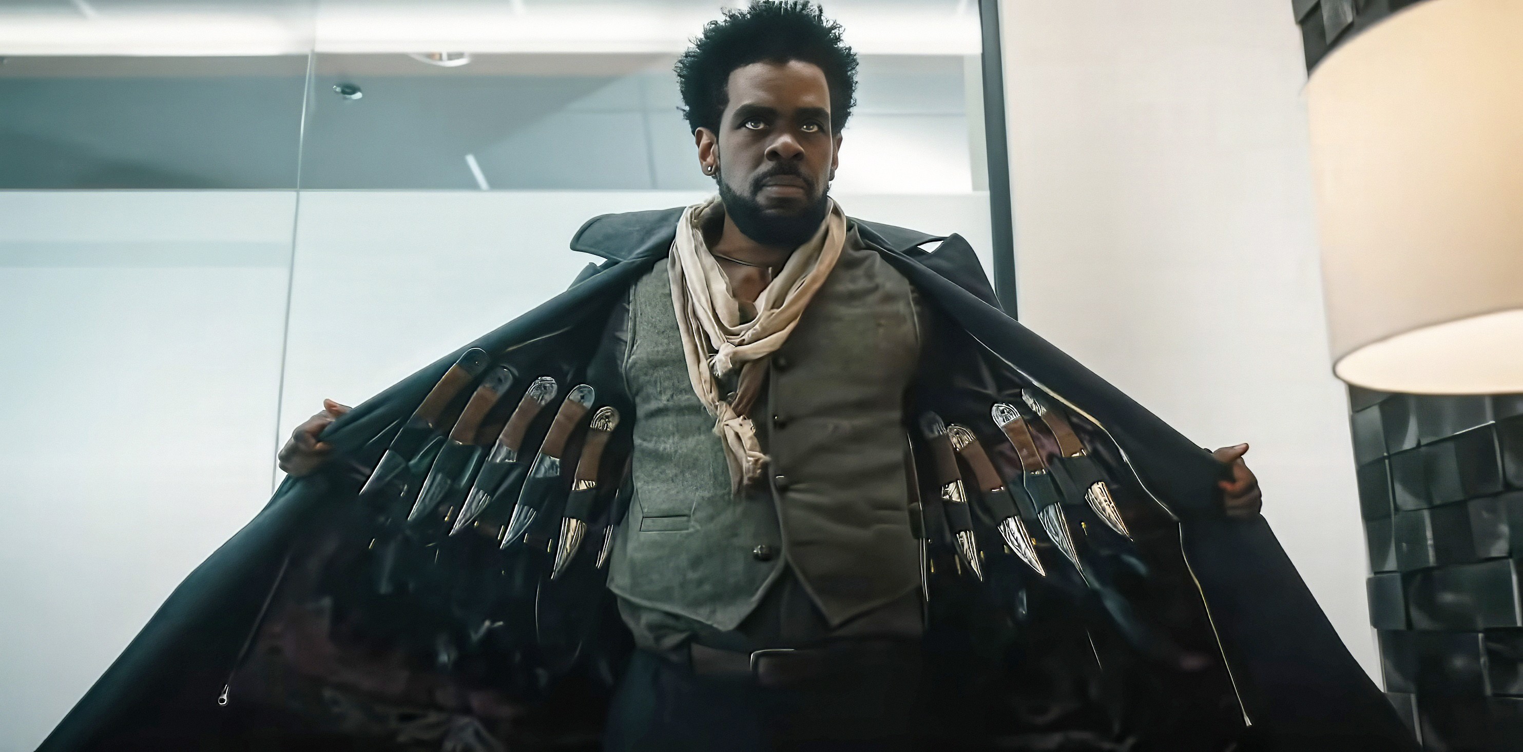 Assassin “The Raven” (Mustafa Shakir), with a grim expression, opens up his trenchcoat to reveal rows of knives in Love Hurts
