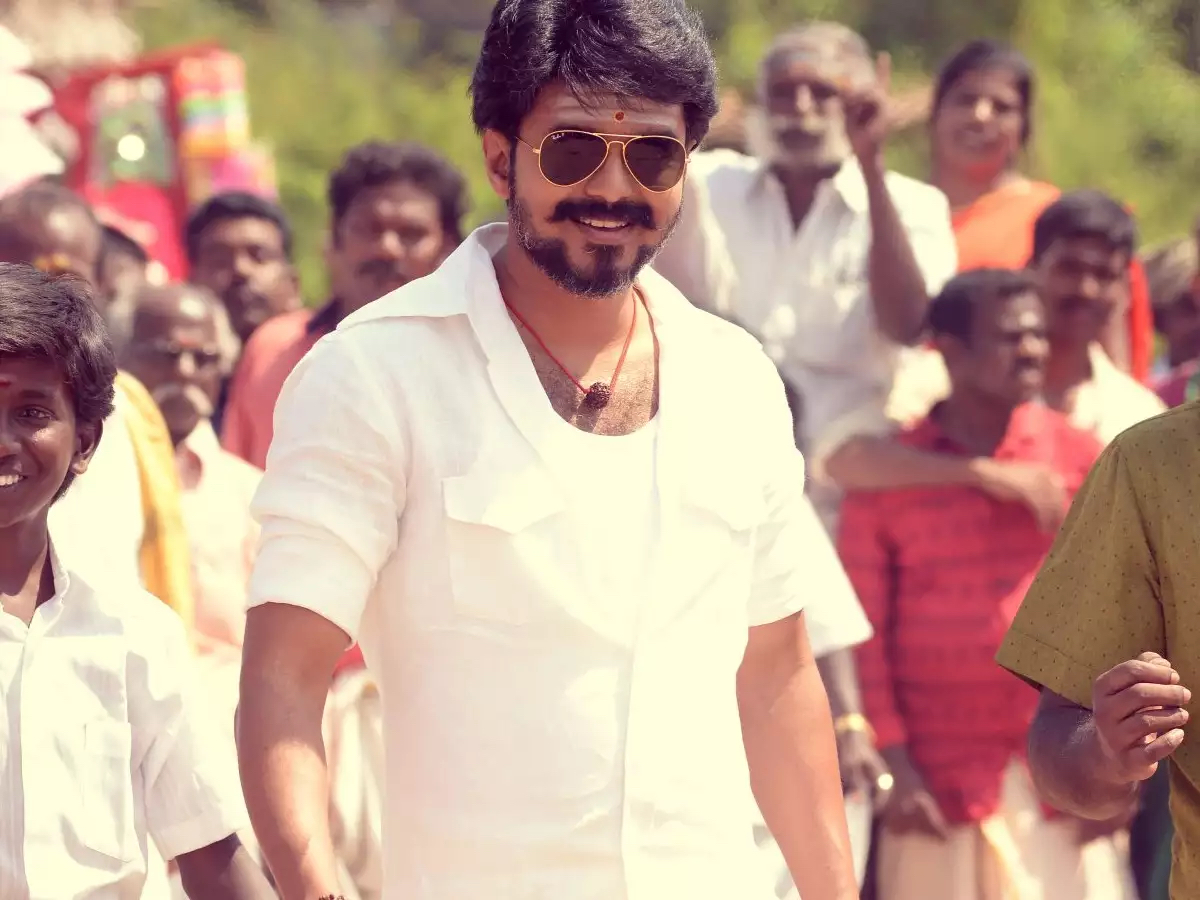 Vijay wears a short-sleeve button-up white shirt, with a white undershirt and a necklace. He also wears sunglasses and has a goatee. His forehead sports a tilak and he is surrounded by a crowd, in Mersal. Vijay wears a short-sleeve button-up white shirt, with a white undershirt and a necklace. He also wears sunglasses and has a goatee. His forehead sports a tilak and he is surrounded by a crowd, in Mersal.