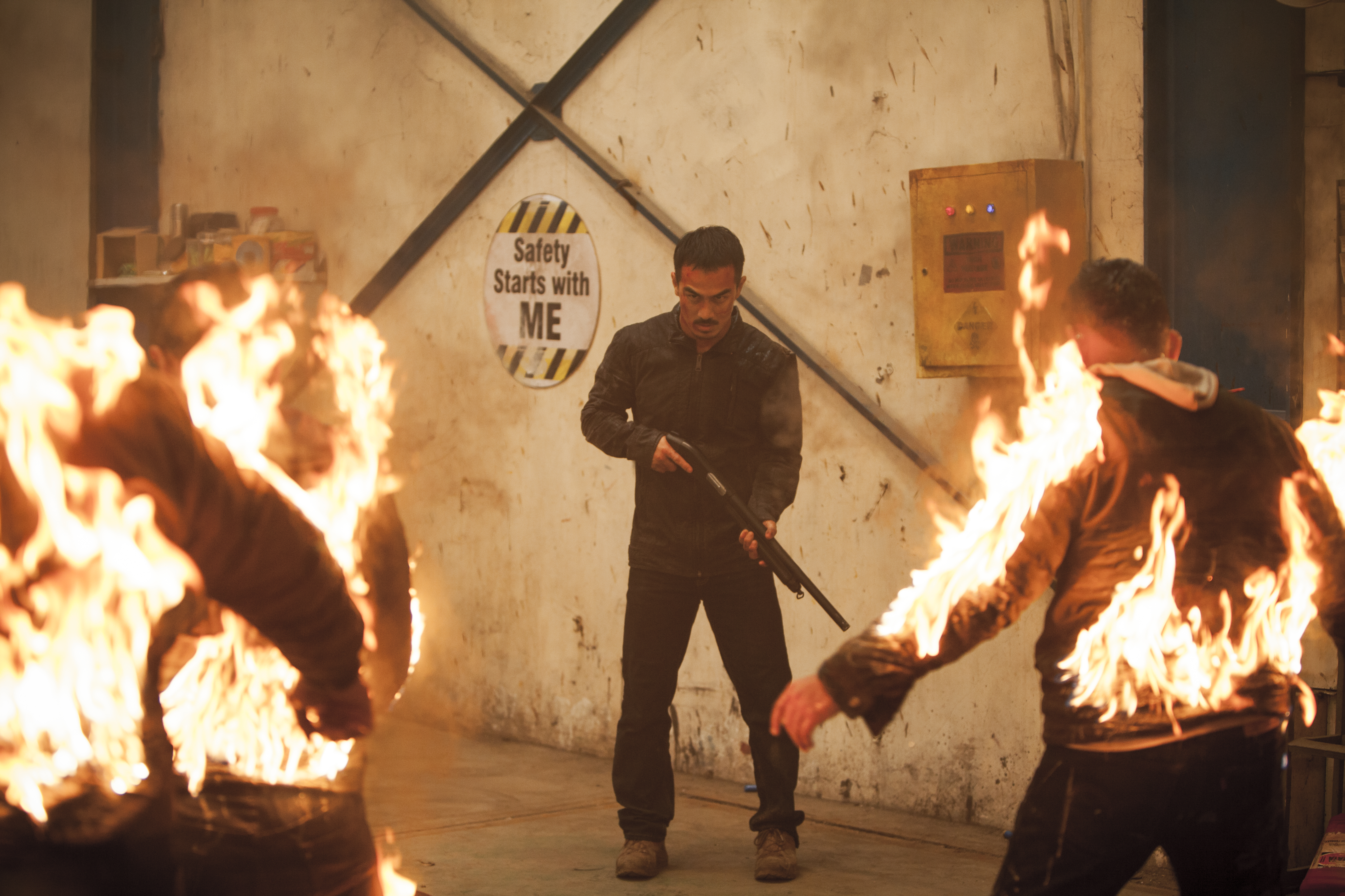 Joe Taslim stands in front of a “Safety starts with me” sign toting a shotgun facing several men on fire in The Night Comes for Us. Joe Taslim stands in front of a “Safety starts with me” sign toting a shotgun facing several men on fire in The Night Comes for Us.
