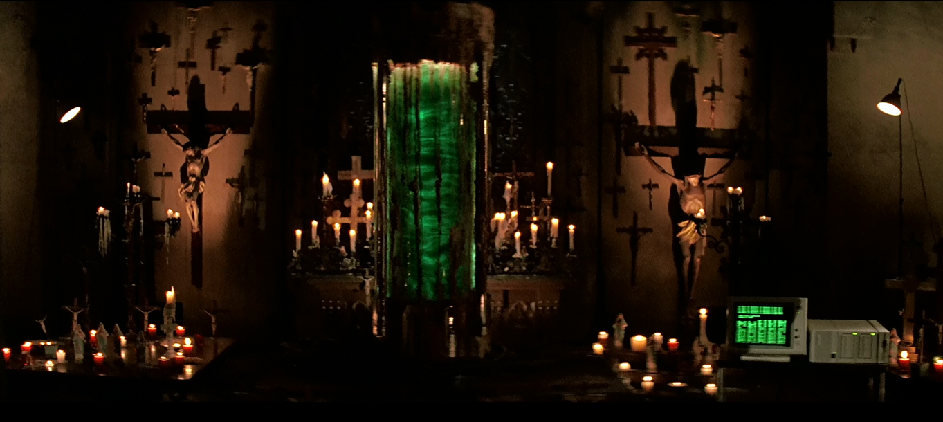 An eerie glowing cylinder filled with green liquid flanked by a wall of crucifixes and computer equipment in Prince of Darkness. An eerie glowing cylinder filled with green liquid flanked by a wall of crucifixes and computer equipment in Prince of Darkness.