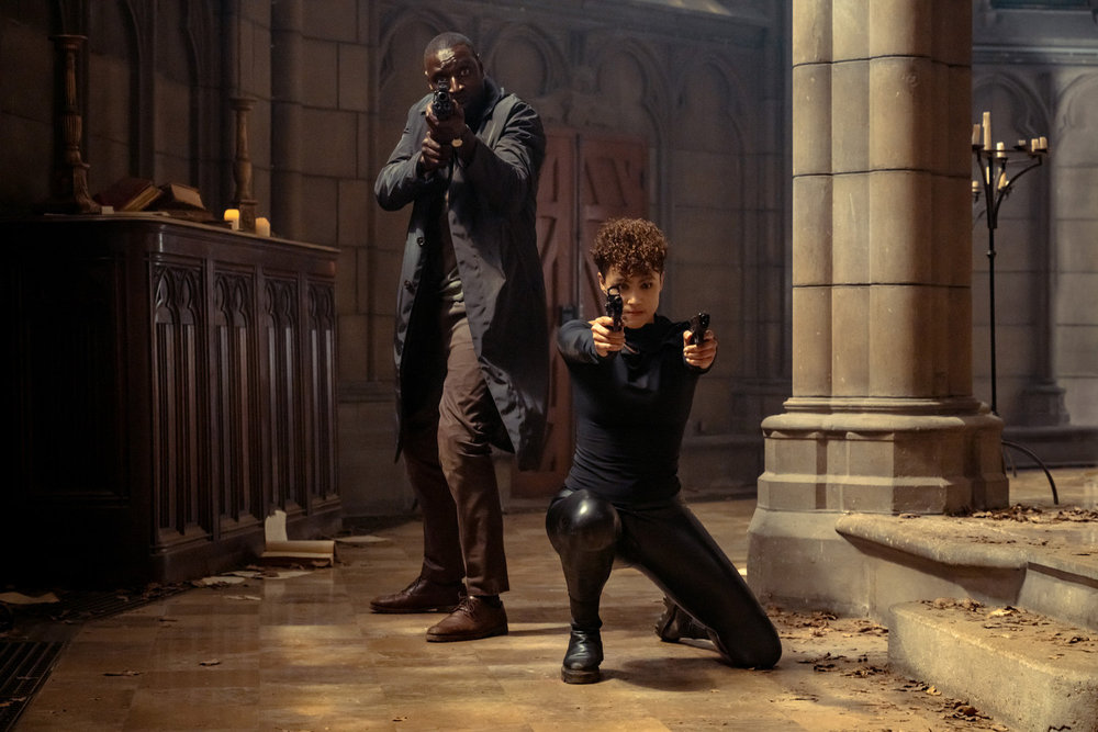 Omar Sy and Nathalie Emmanuel posing with a rifle and pistols in a church in The Killer. Omar Sy and Nathalie Emmanuel posing with a rifle and pistols in a church in The Killer.