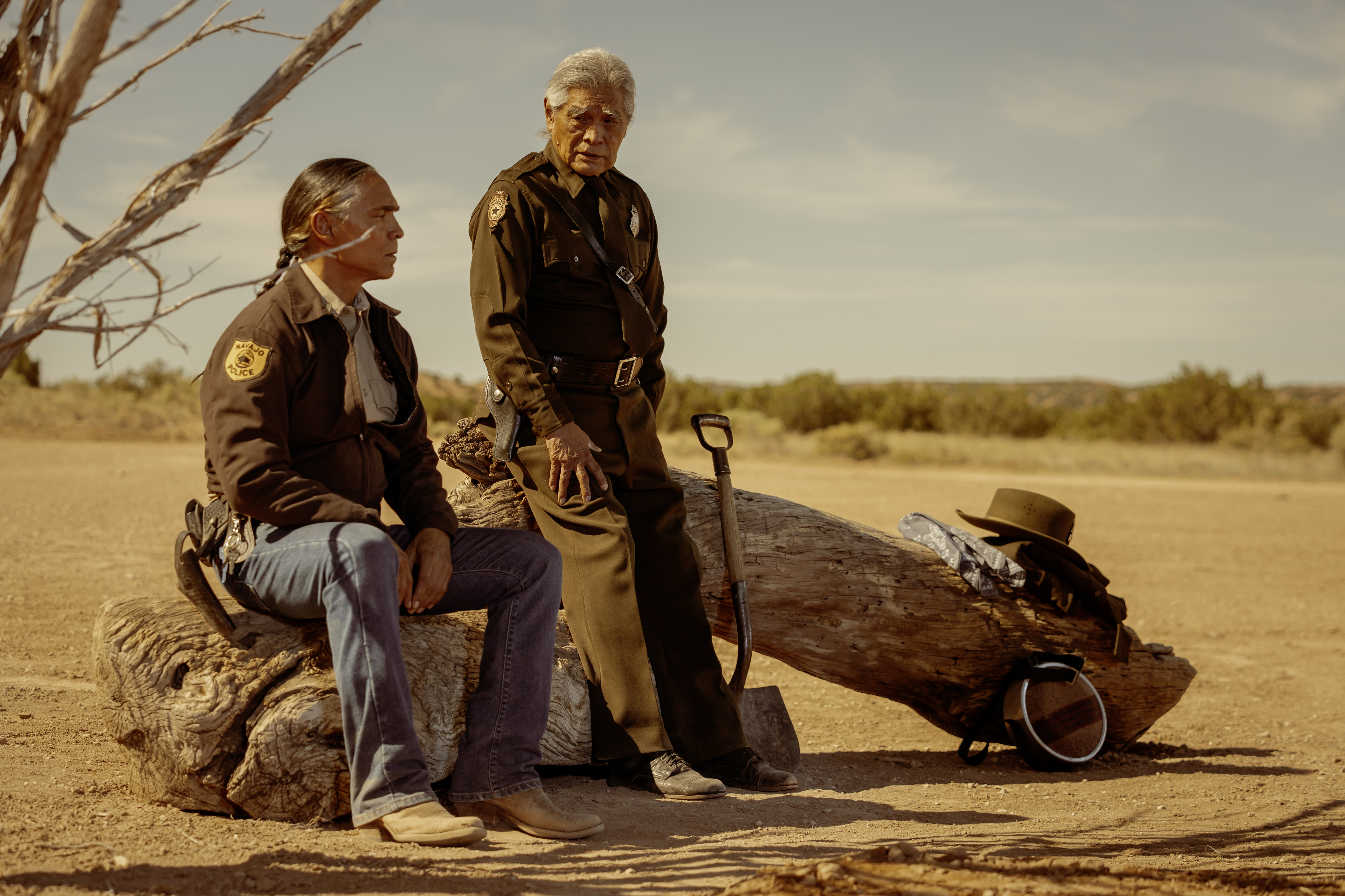 Two sheriffs sit on a log with a shovel and some supplies in a still from Dark Winds season 3 Two sheriffs sit on a log with a shovel and some supplies in a still from Dark Winds season 3