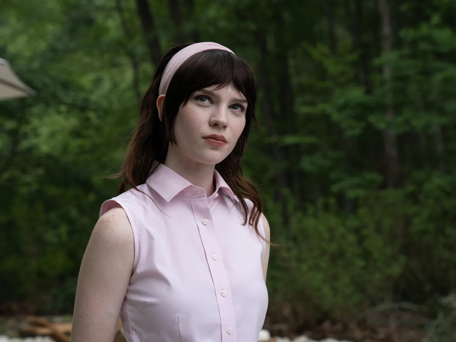 A brown haired woman in a short-sleeved, pink buttoned-up shirt and matching headband standing against the backdrop of a forest in Companion. A brown haired woman in a short-sleeved, pink buttoned-up shirt and matching headband standing against the backdrop of a forest in Companion.