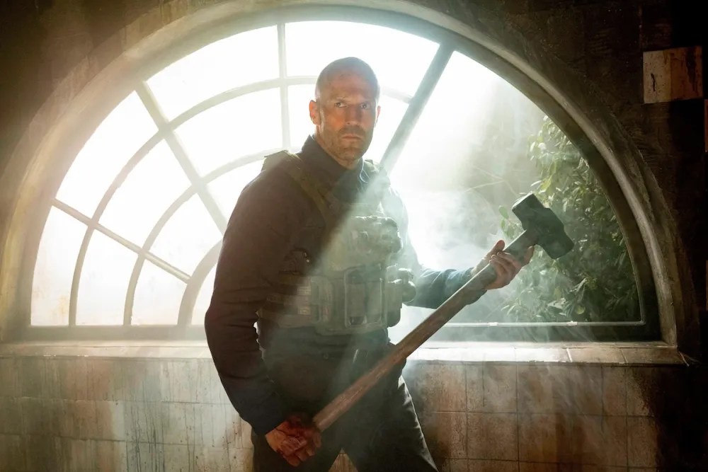 Jason Statham wearing an armored vest and holding a sledgehammer in front of a sunny half-circle window in A Working Man. Jason Statham wearing an armored vest and holding a sledgehammer in front of a sunny half-circle window in A Working Man.