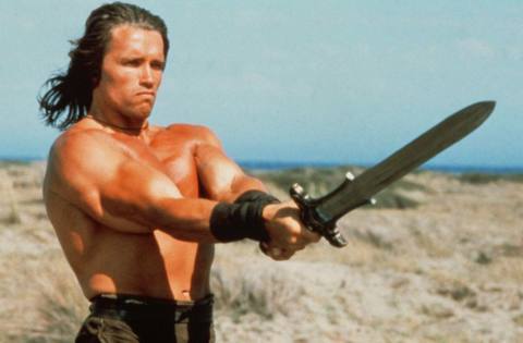Arnold Schwarzenegger as Conan the Barbarian holding a sword while not wearing a shirt
