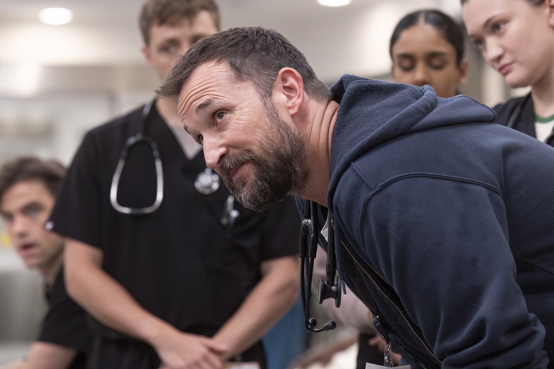Noah Wyle, surrounded by student doctors, looks up from a hunched position in The Pitt Noah Wyle, surrounded by student doctors, looks up from a hunched position in The Pitt