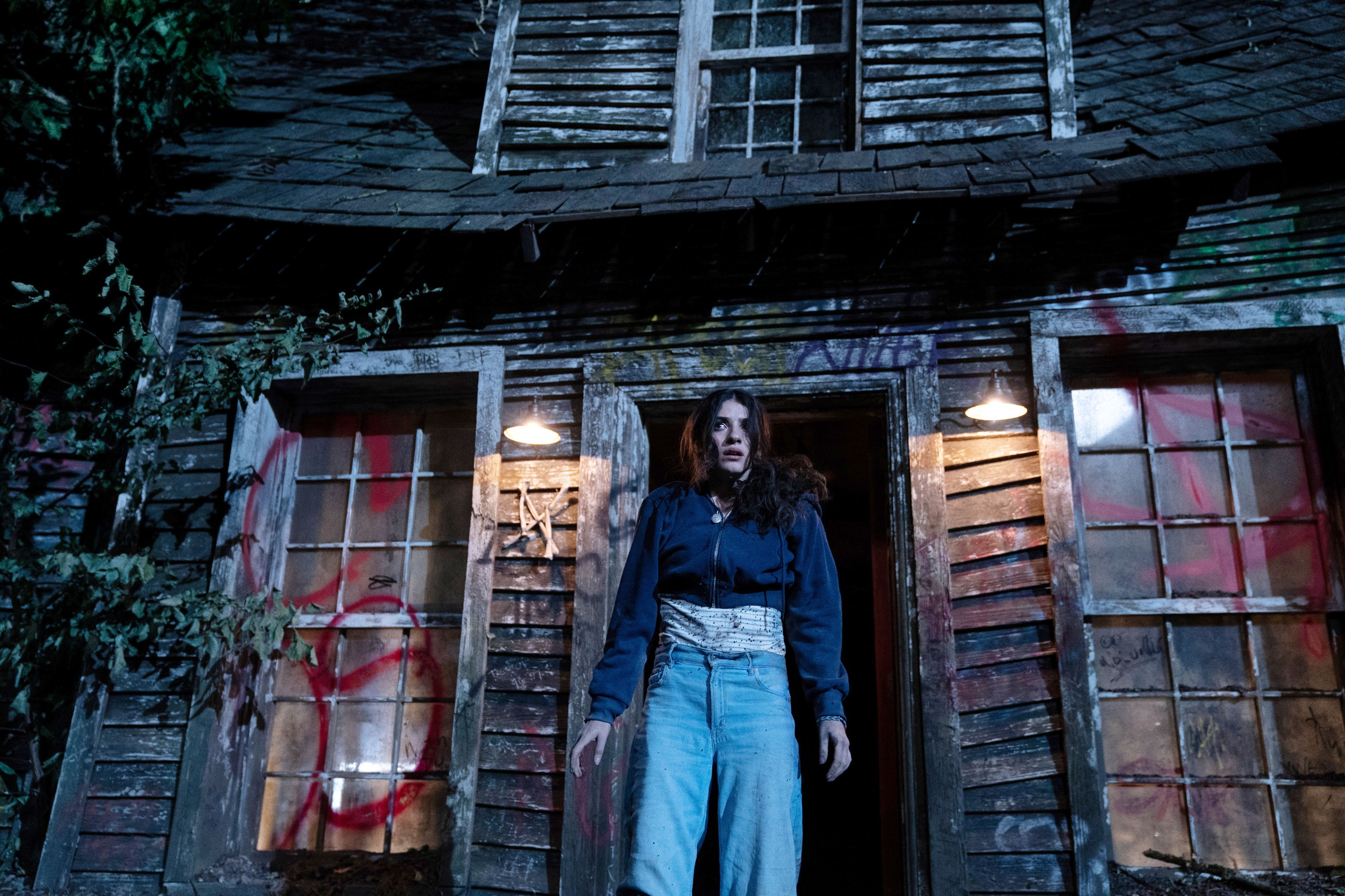 Clover (Ella Rubin), a teenager in a high-waisted dark blue sweater and light blue pants, stands in the front doorway of a battered-looking house covered with red spray-paint graffiti in Until Dawn Clover (Ella Rubin), a teenager in a high-waisted dark blue sweater and light blue pants, stands in the front doorway of a battered-looking house covered with red spray-paint graffiti in Until Dawn