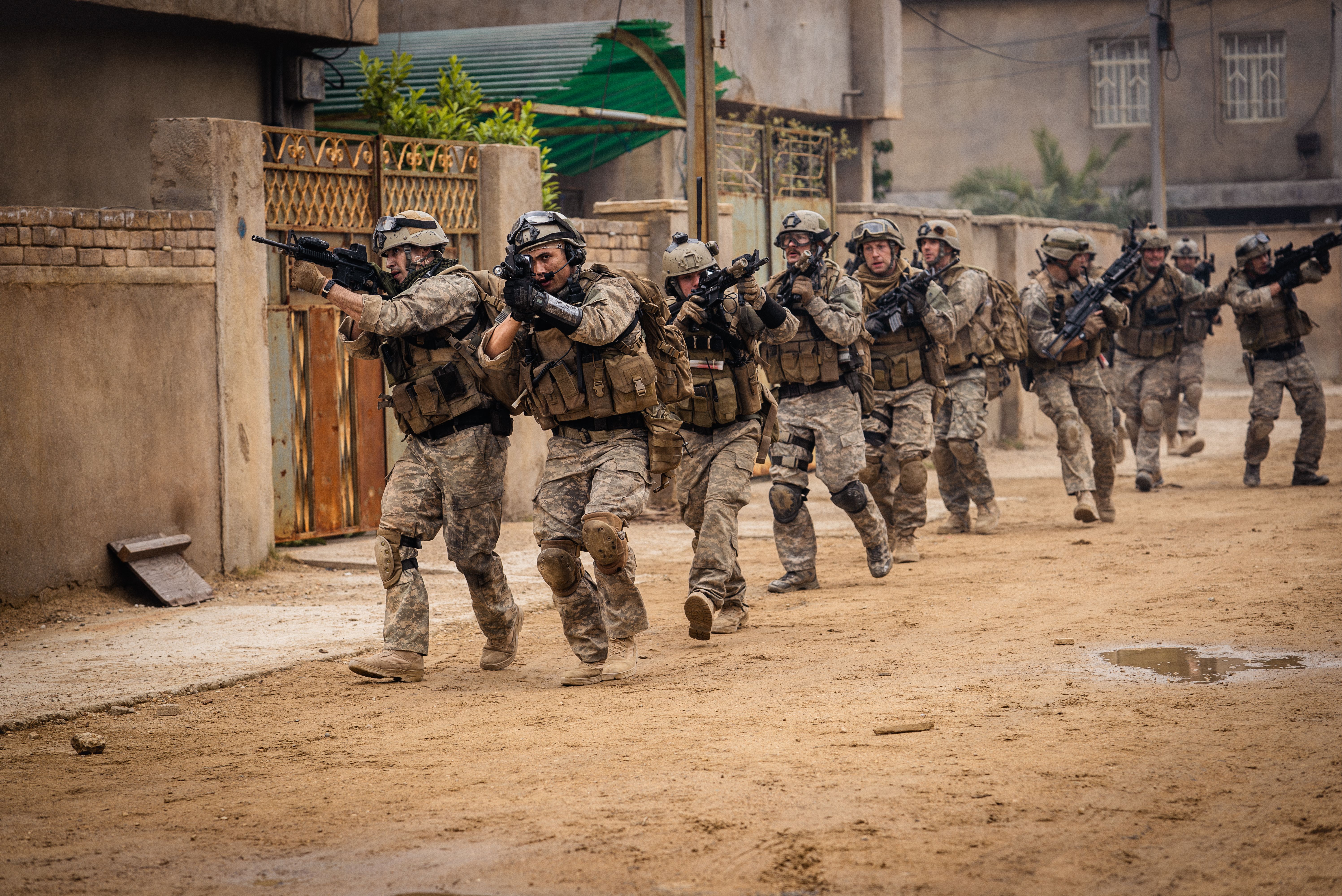 Several Navy SEALs walk through a sandy, unpaved Iraq street with their weapons held up in the movie Warfare Several Navy SEALs walk through a sandy, unpaved Iraq street with their weapons held up in the movie Warfare