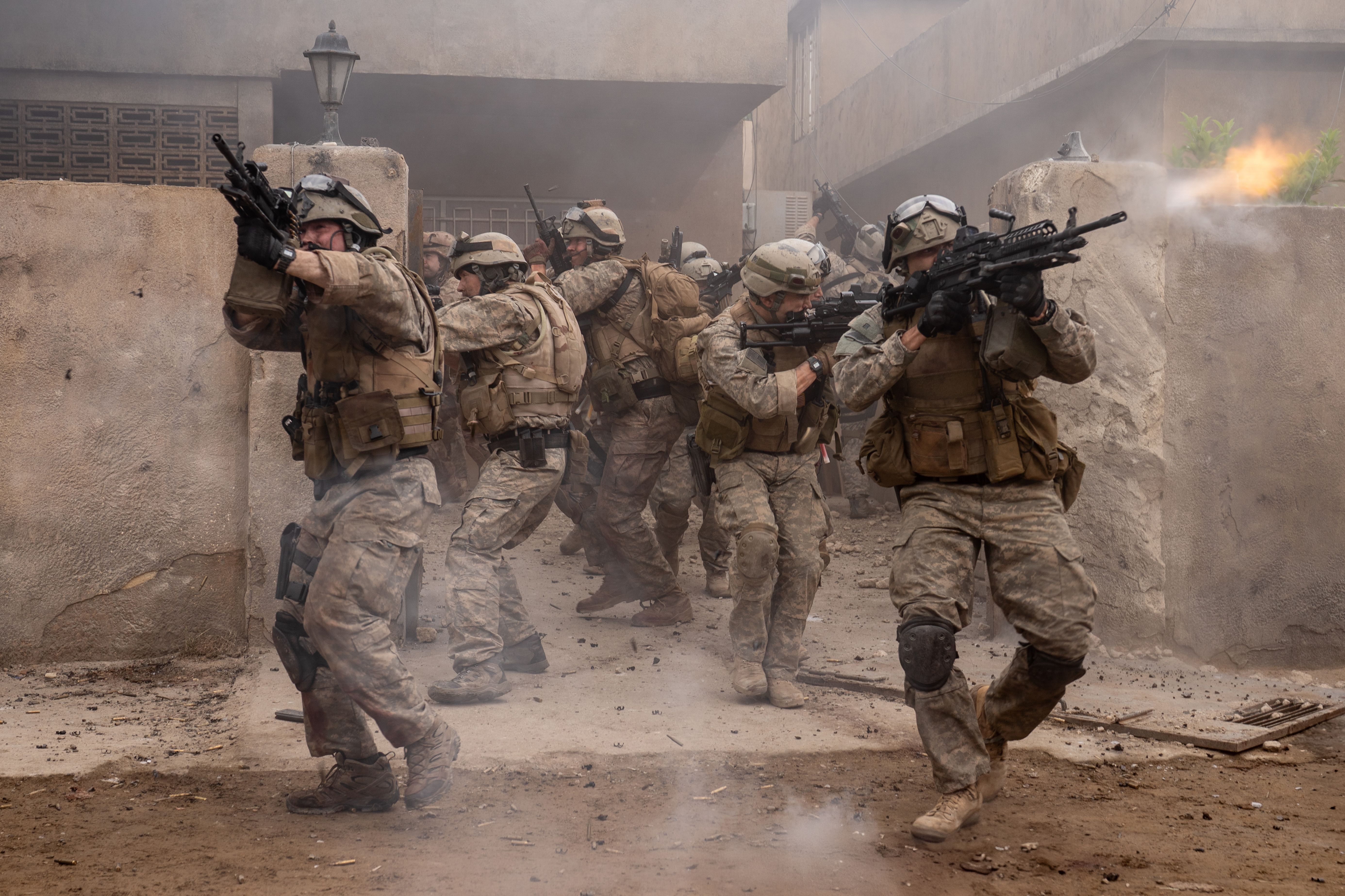 Several Navy SEALs walk out of a gate firing assault rifles in the movie Warfare Several Navy SEALs walk out of a gate firing assault rifles in the movie Warfare