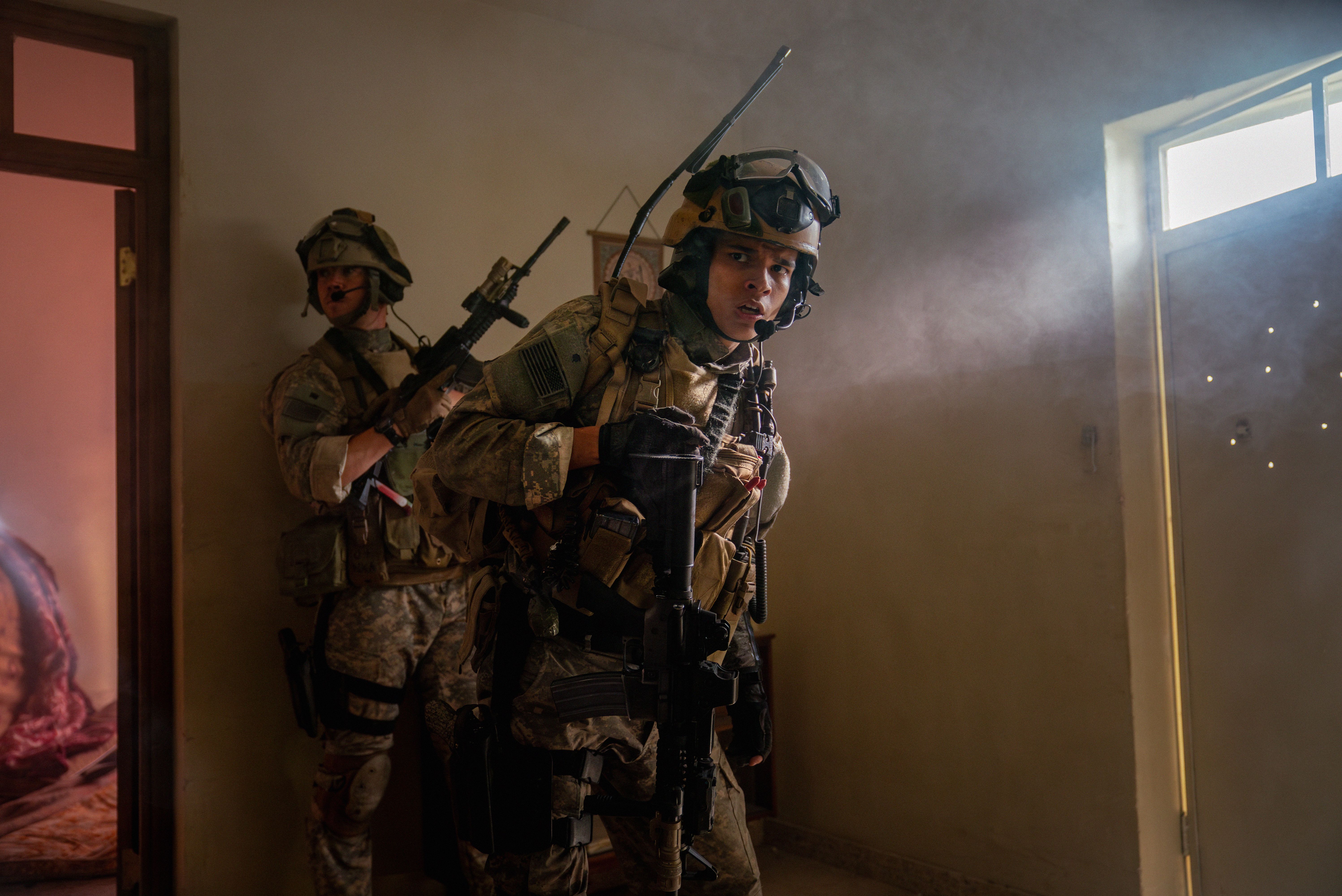 Two soldiers in the movie Warfare hold weapons while standing near a door with bullet holes in it Two soldiers in the movie Warfare hold weapons while standing near a door with bullet holes in it