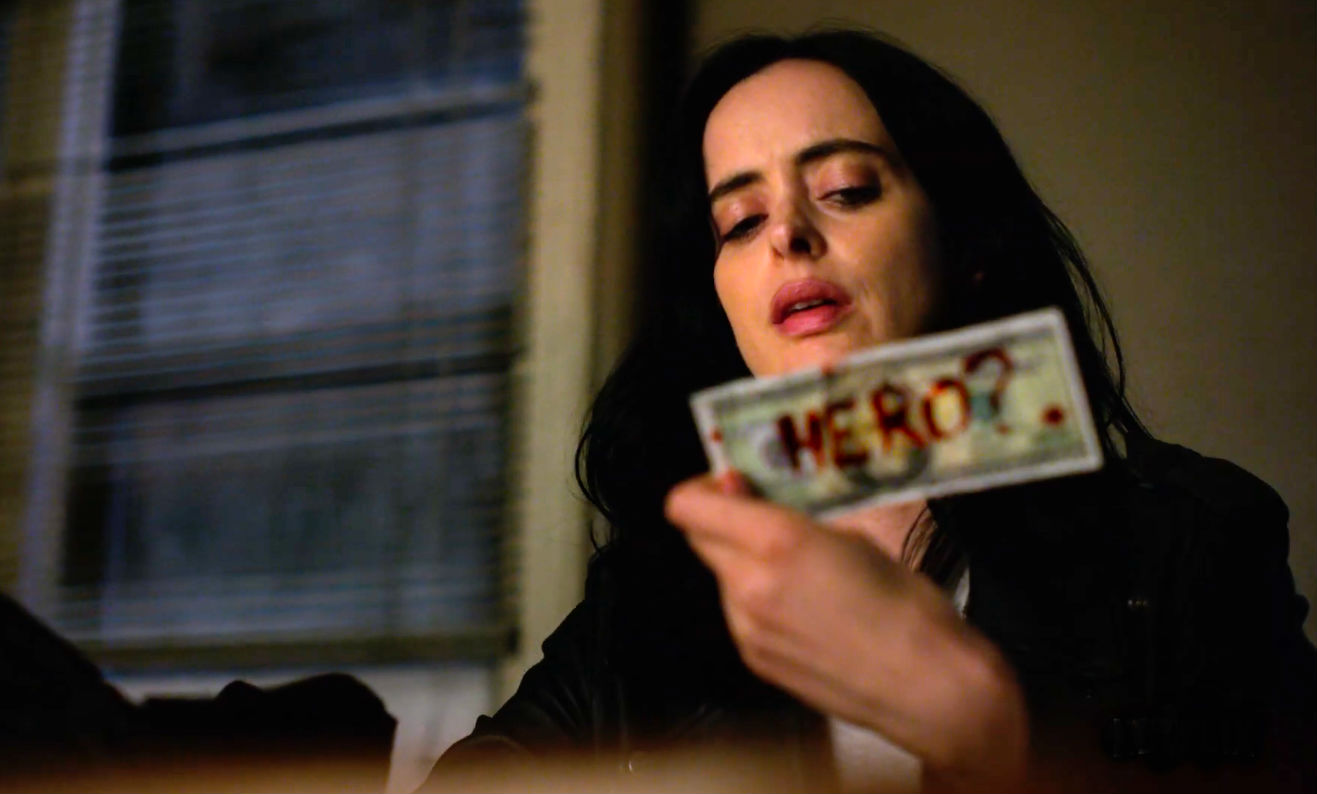 A screenshot of Jessica Jones from the Netflix series holding up a dollar bill onto which someone has scrawled the word “Hero?”