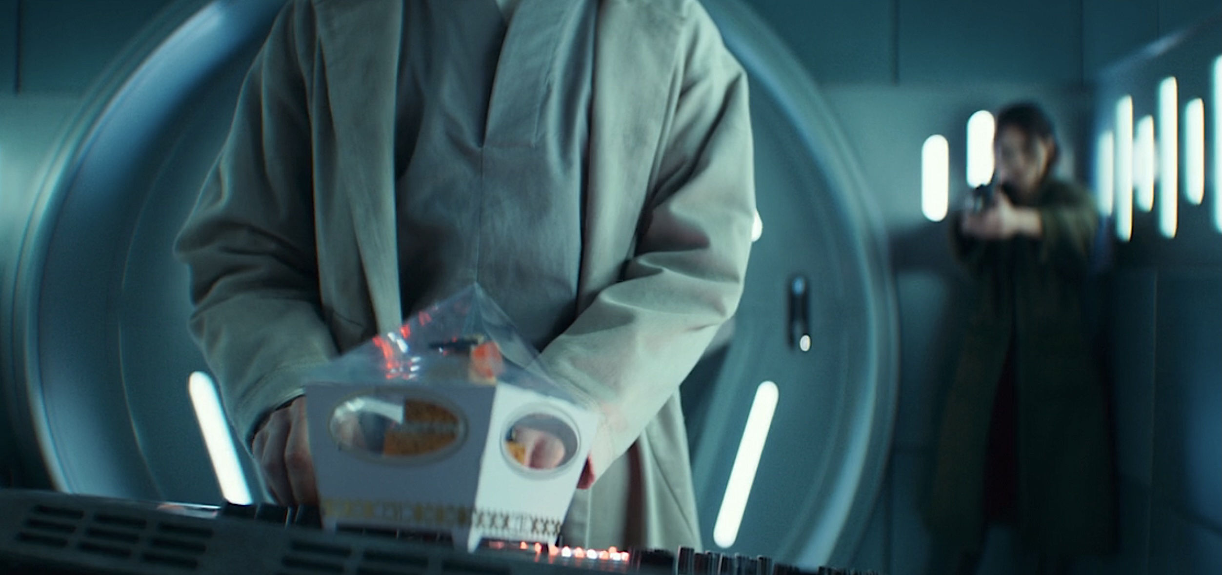 A shot from season 2, episode 6 of Andor, with Bix (Adria Arjona) in the background, out of focus, pointing a blaster at Imperial torturer Doctor Gorst (Joshua James), whose back is to her. He’s standing in the foreground at a computer panel, having just put down a fancy cupcake in a decorative box on the panel in front of him, in the extreme foreground. A shot from season 2, episode 6 of Andor, with Bix (Adria Arjona) in the background, out of focus, pointing a blaster at Imperial torturer Doctor Gorst (Joshua James), whose back is to her. He’s standing in the foreground at a computer panel, having just put down a fancy cupcake in a decorative box on the panel in front of him, in the extreme foreground.