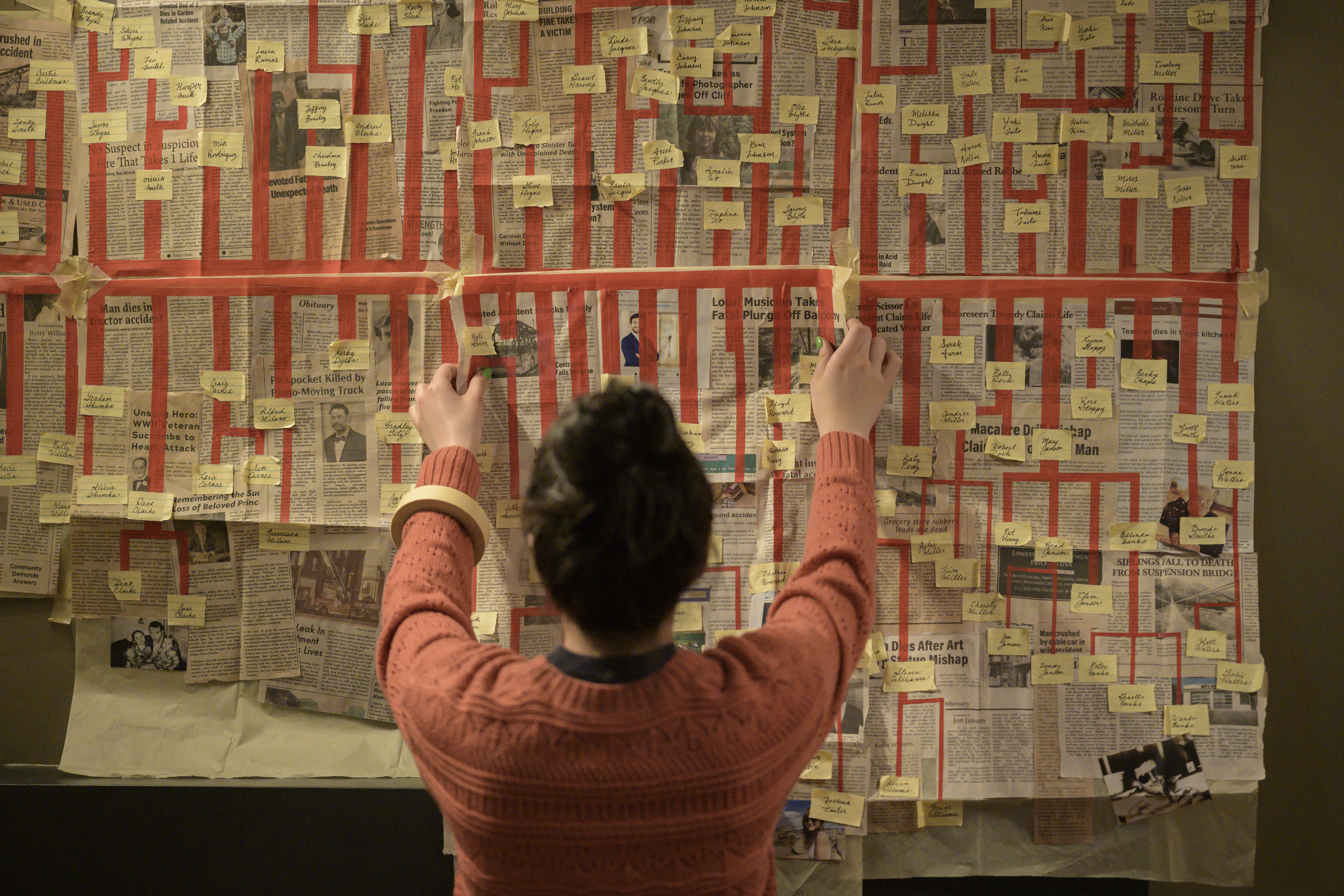Stefanie (Kaitlyn Santa Juana), a dark-haired young woman, stands with her back to the camera, in front of an elaborate timeline on the wall built on newspaper clips covered with red ink lines and post-its, in Final Destination: Bloodlines