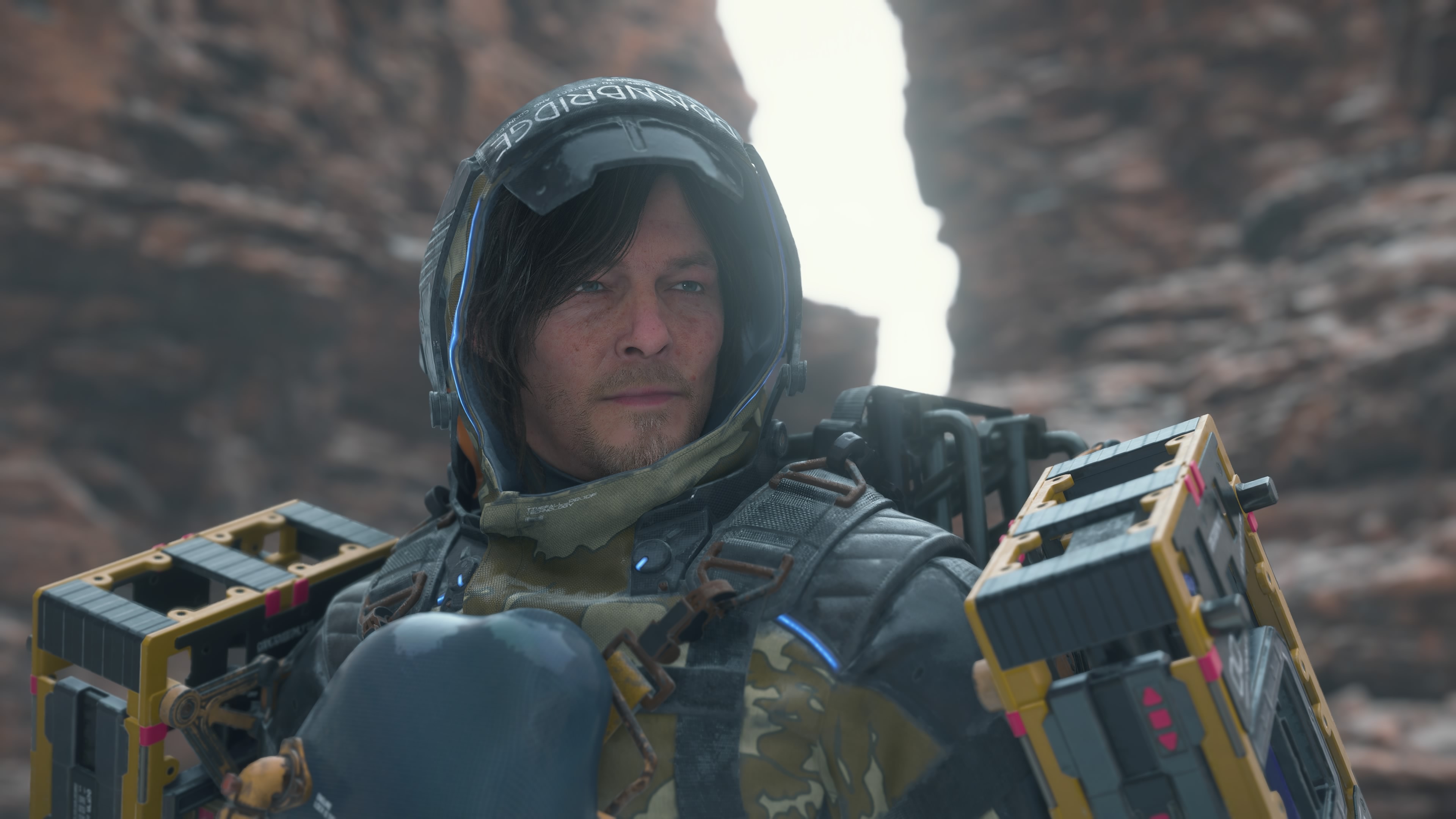 Death Stranding 2 stats: Sam geared up with cargo and his coat, looking off to the side while standing in a ravine.