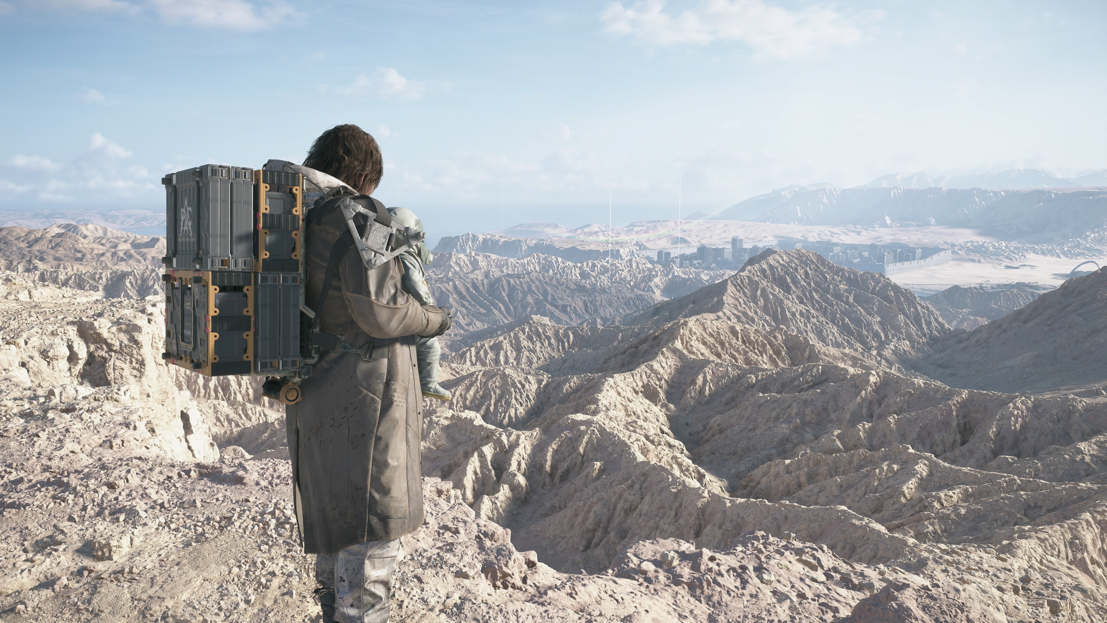 Death Stranding 2 birthday DOOMS: Sam, with Lou on his chest, standing on a mountain overlooking a desert valley.