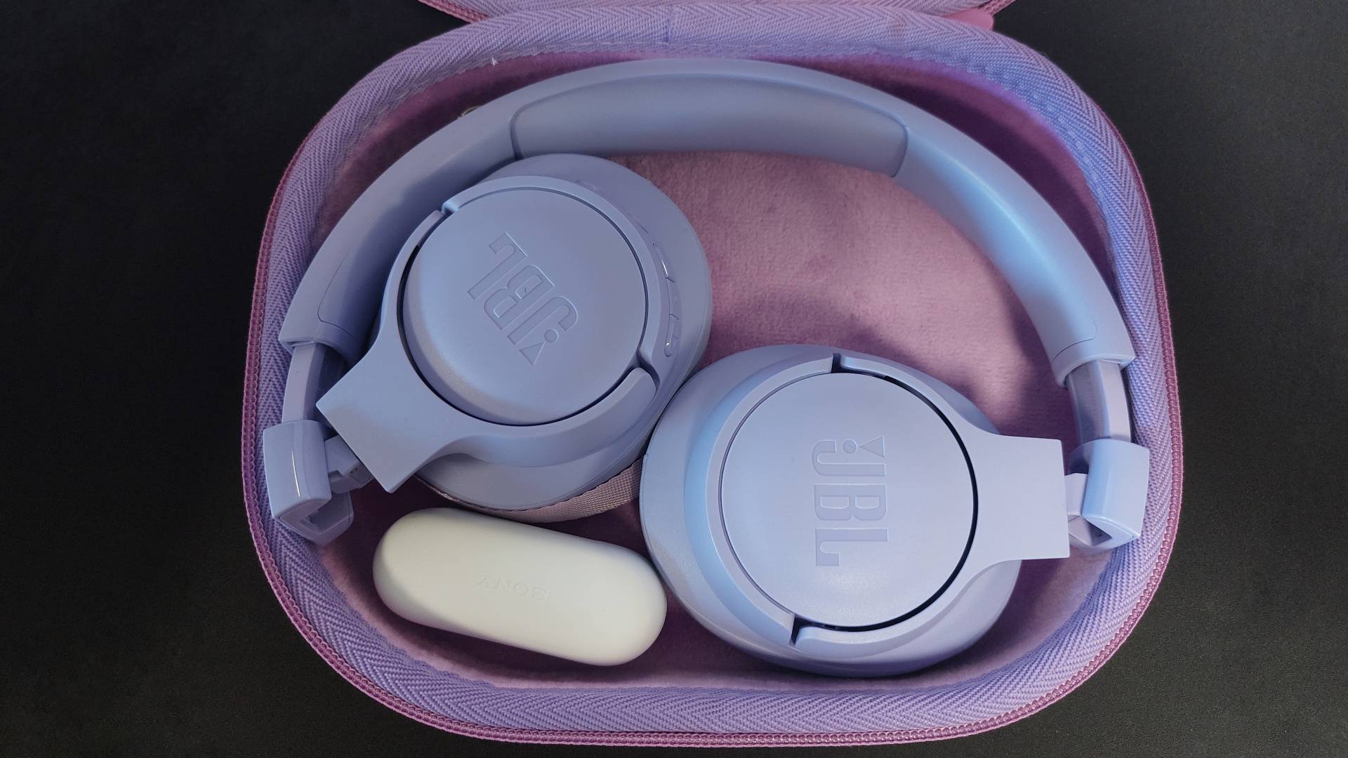 The Sony WF-C510 earbuds on a black desk mat. They are seen here inside their sleek charging case. The case has been placed beside a lilac pair of the JBL TUNE 720BT headphones to show the dramatic size difference.