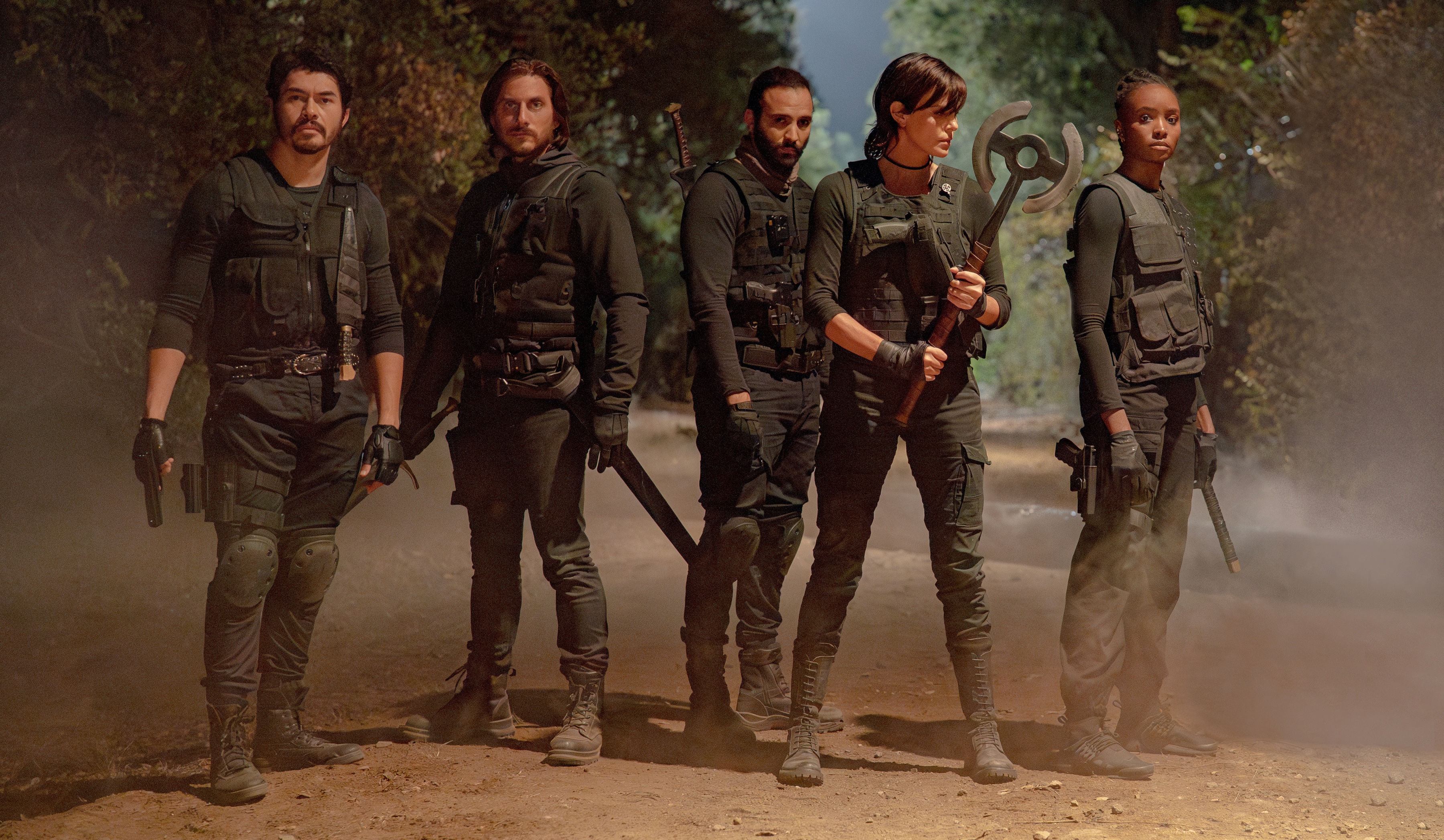 A team of armed immortals and their partners (played by Henry Golding, Luca Marinelli, Marwan Kenzari, Charlize Theron, and Kiki Layne) wearing dark military-style clothing stand on a dusty road in The Old Guard 2 A team of armed immortals and their partners (played by Henry Golding, Luca Marinelli, Marwan Kenzari, Charlize Theron, and Kiki Layne) wearing dark military-style clothing stand on a dusty road in The Old Guard 2