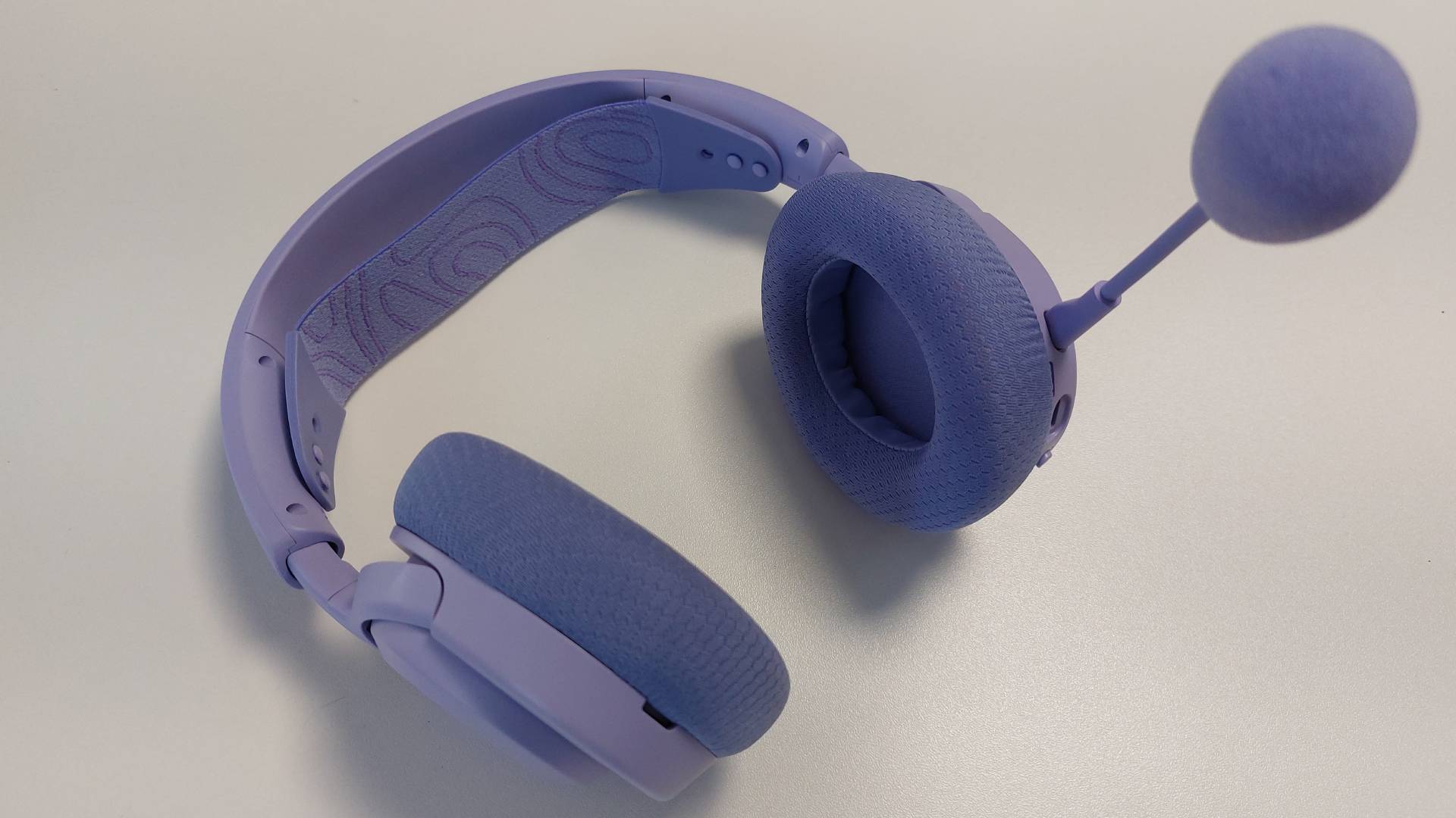 The SteelSeries Arctis Nova 3 X seen on a desk. The detachable mic arm is attached, reaching up towards the viewer. This is the Lavender colourway.